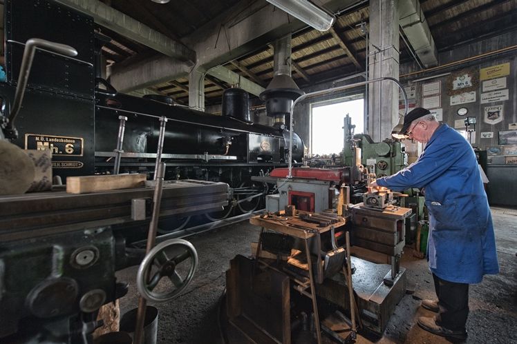 Ein Mann arbeitet in einer Werkstatt an einer Maschine neben einer alten Dampflokomotive.