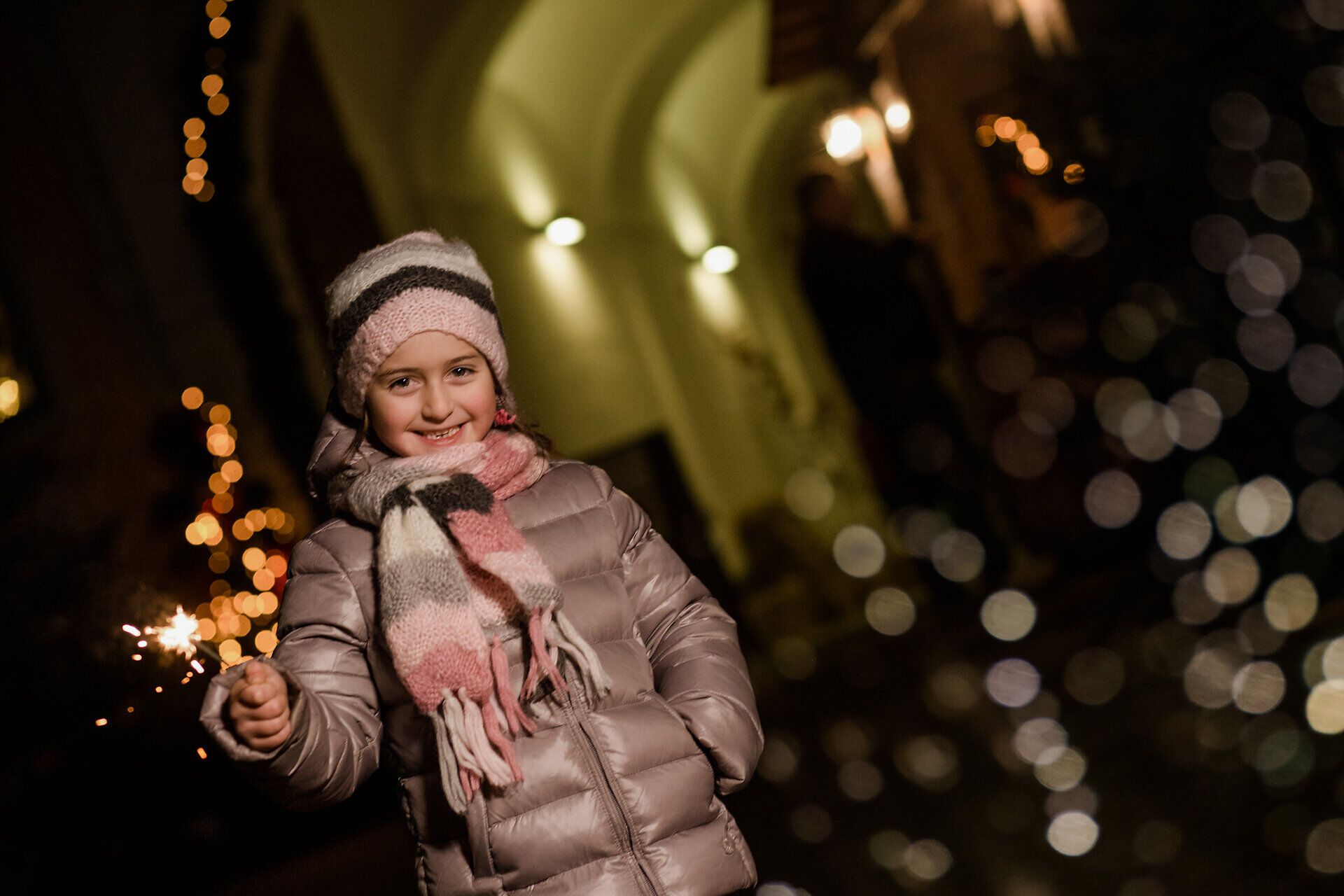 Ein strahlendes Lächeln erhellt das Gesicht eines kleinen Mädchens, während sie mit einem funkelnden Wunderkerze in der Hand die festliche Atmosphäre genießt. Umgeben von glitzernden Lichtern und dem Duft von gebrannten Mandeln, wird die Vorfreude auf das Weihnachtsfest spürbar.
