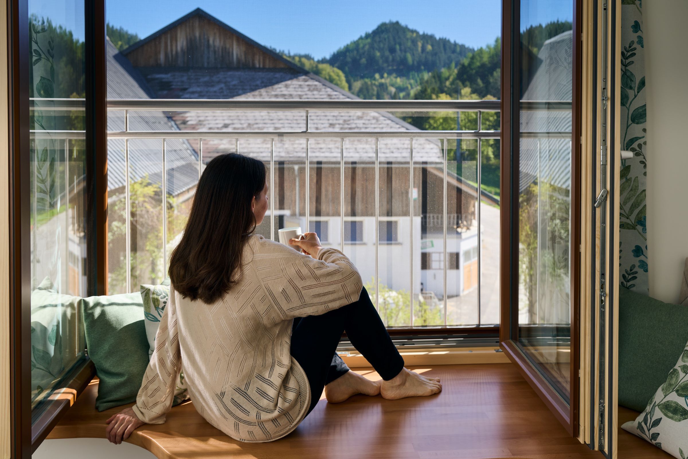 Eine Frau sitzt auf einer Fensterbank und blickt auf eine ländliche Landschaft mit Bergen im Hintergrund.