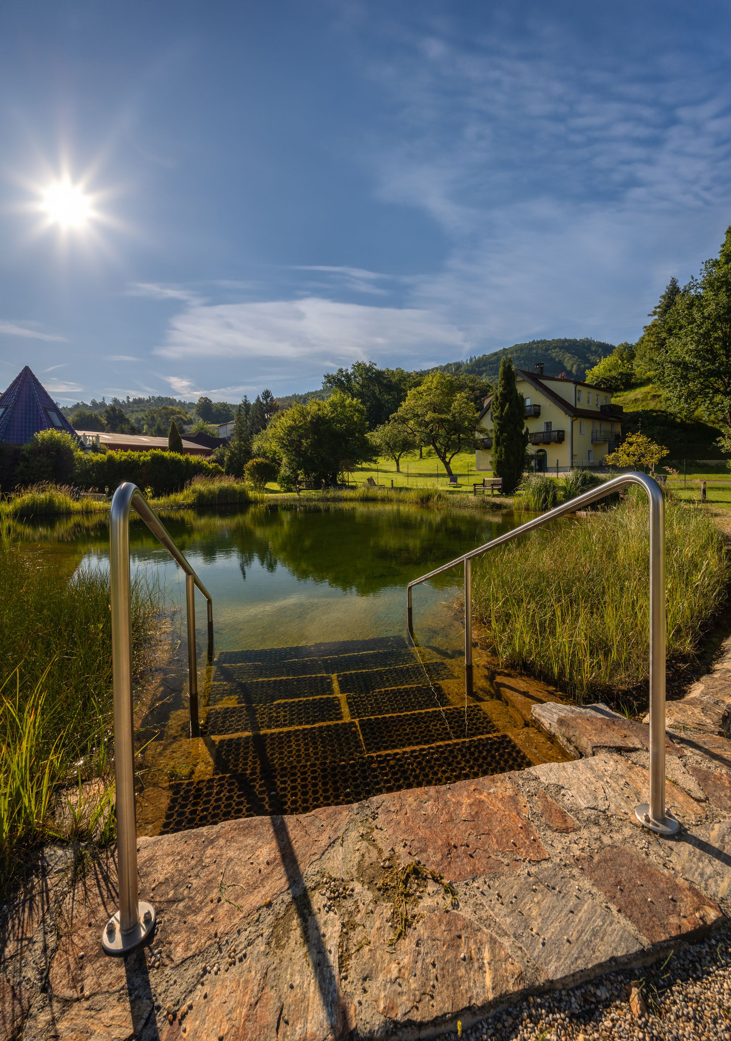 Treppen führen in einen Teich mit klarem Wasser, umgeben von grüner Landschaft und einem Haus im Hintergrund, unter einem strahlend blauen Himmel mit Sonne.