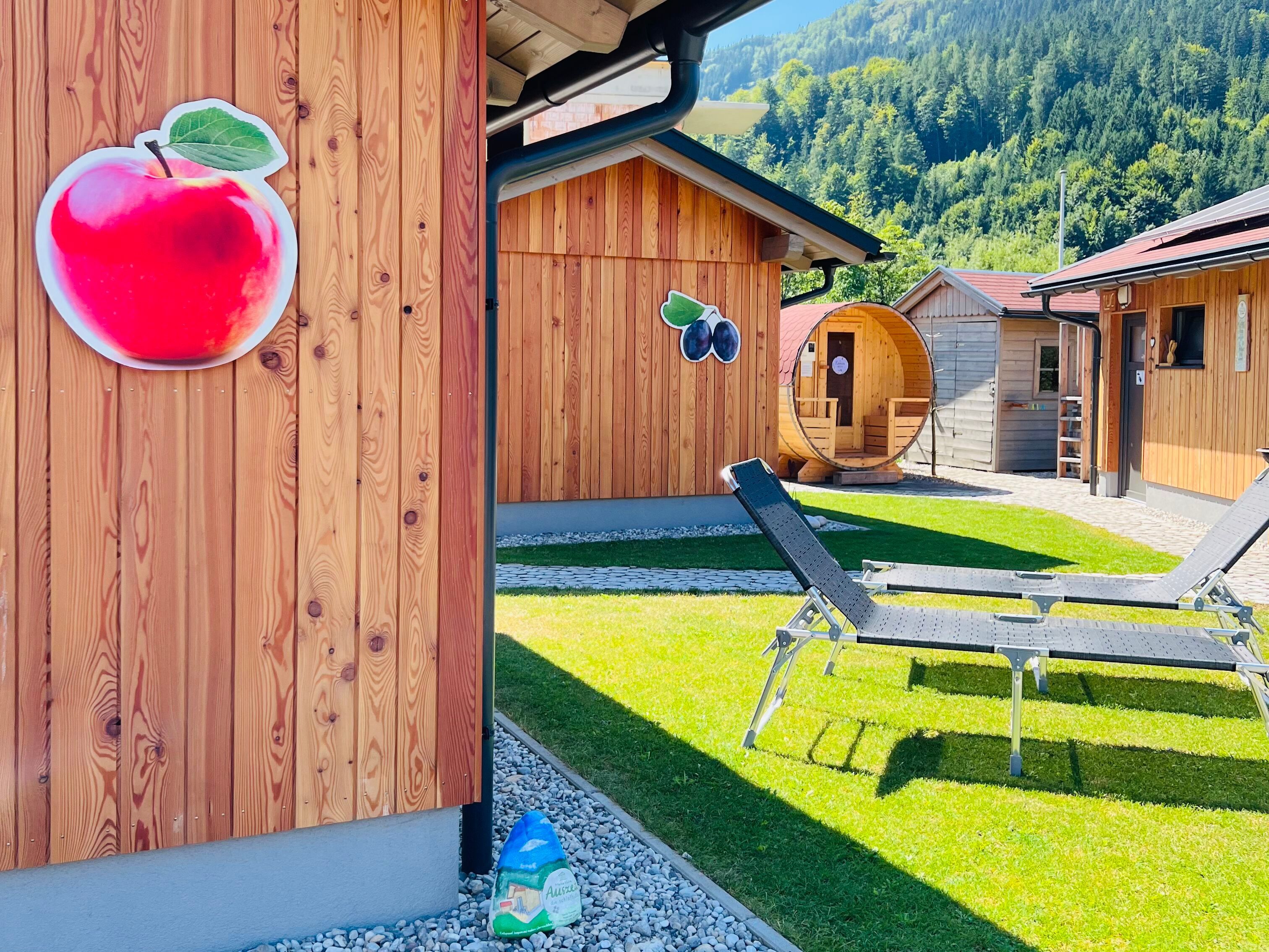 Holzhütten mit Obstdekoration und Liegestühlen auf einer Wiese, umgeben von Bergen.