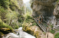 Ein malerischer Wanderweg schlängelt sich entlang des rauschenden Wassers, umgeben von üppigem Grün und majestätischen Felsen. Familien genießen die frische Luft und die atemberaubenden Ausblicke, während sie die Natur in vollen Zügen erleben.