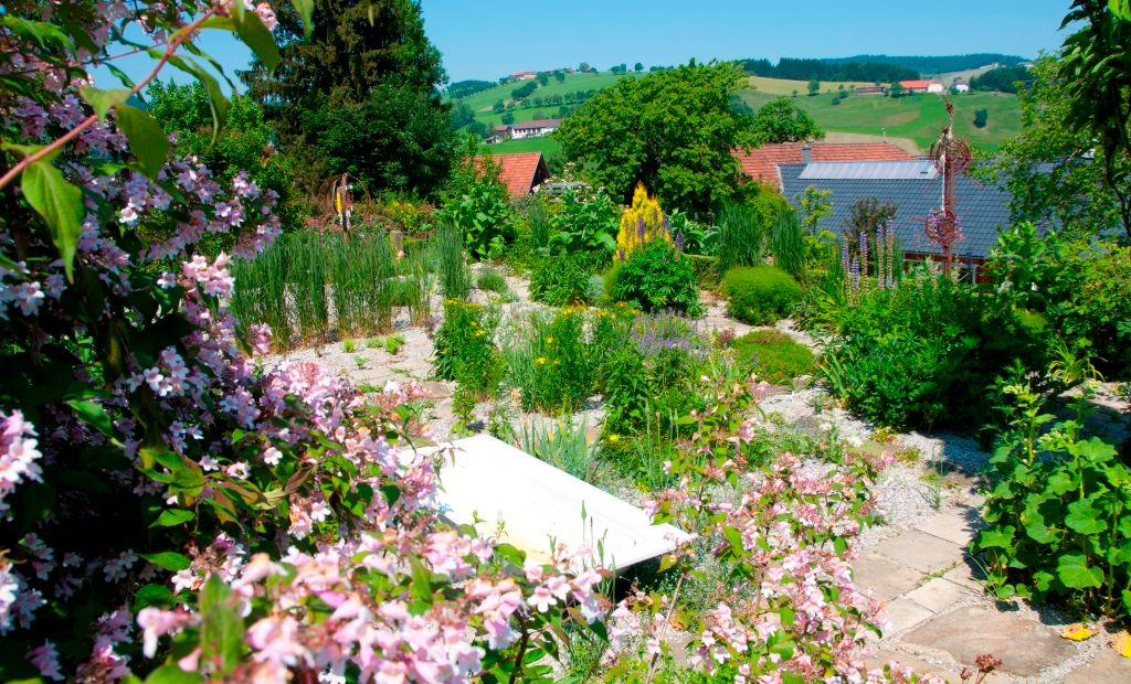 Ein blühender Garten mit bunten Blumen und grünen Pflanzen, im Hintergrund Hügel und ein Hausdach.