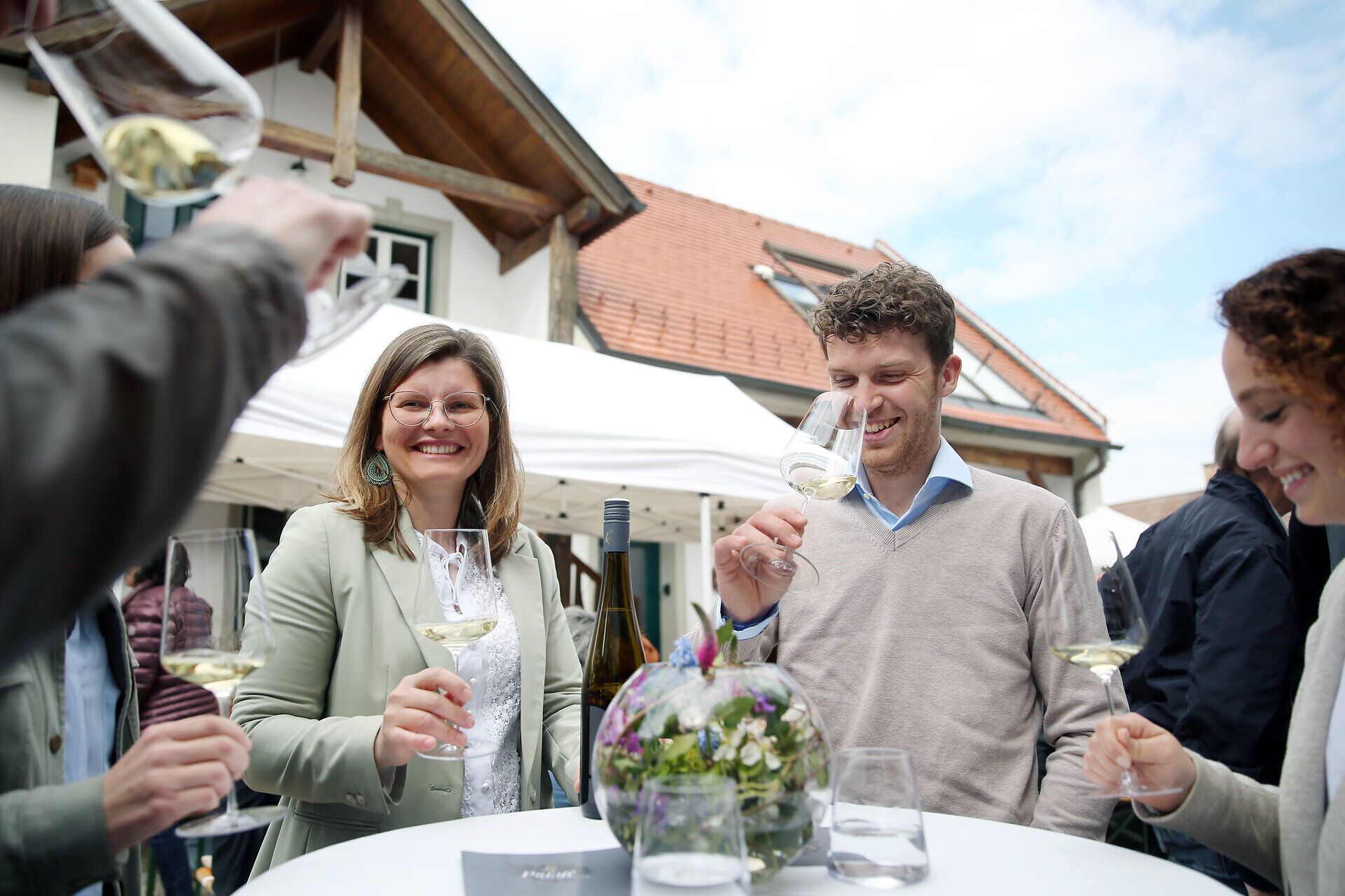 Inmitten einer festlichen Atmosphäre genießen die Gäste erlesene Weine und kulinarische Köstlichkeiten. Die fröhlichen Gesichter und das Anstoßen mit Gläsern schaffen eine einladende Stimmung, die zum Verweilen einlädt.