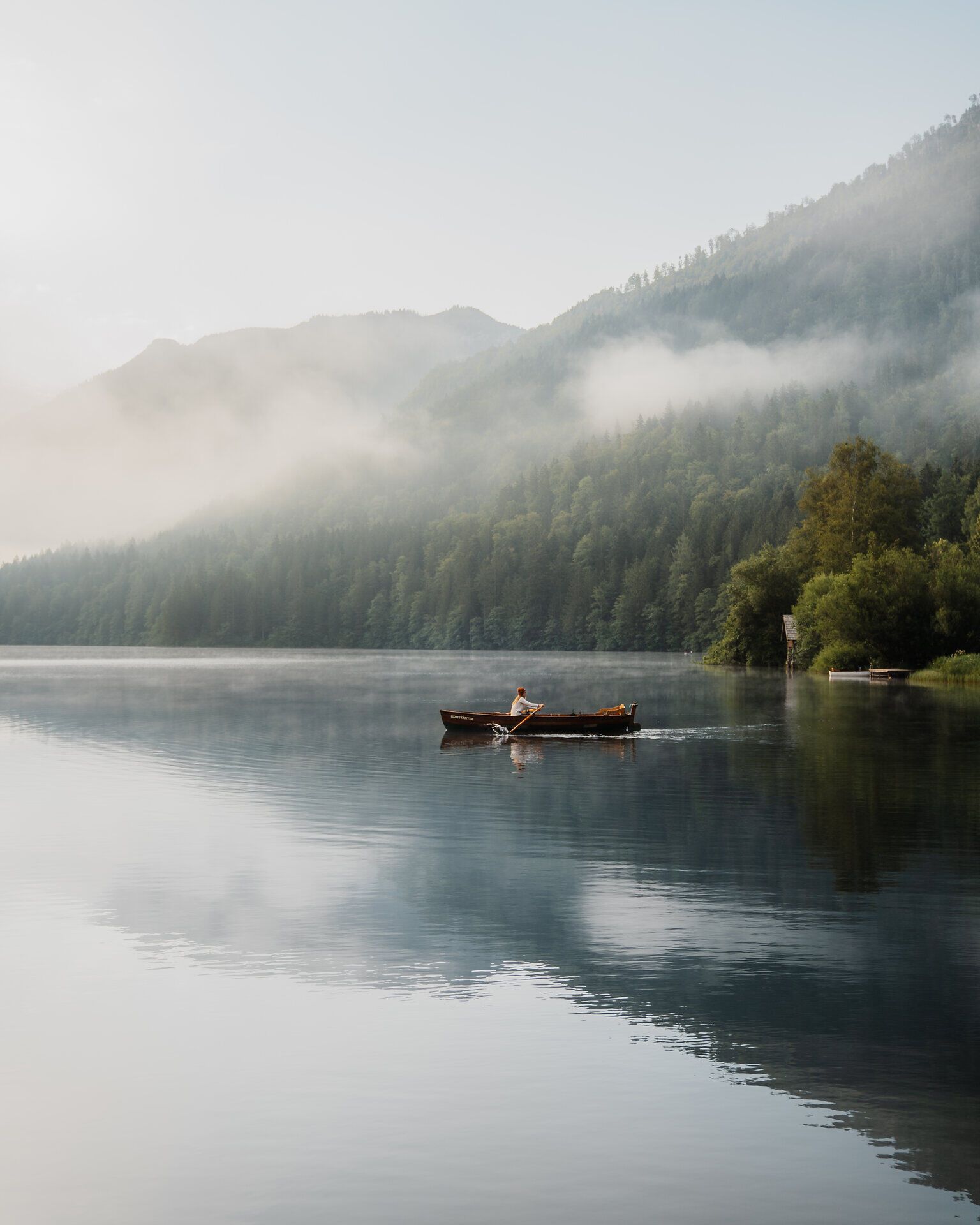 Ein ruhiger Morgen am See, wo sanfte Nebelschwaden über das Wasser gleiten. Ein einzelnes Boot schneidet durch die spiegelglatte Oberfläche und lädt dazu ein, die friedliche Natur zu genießen.
