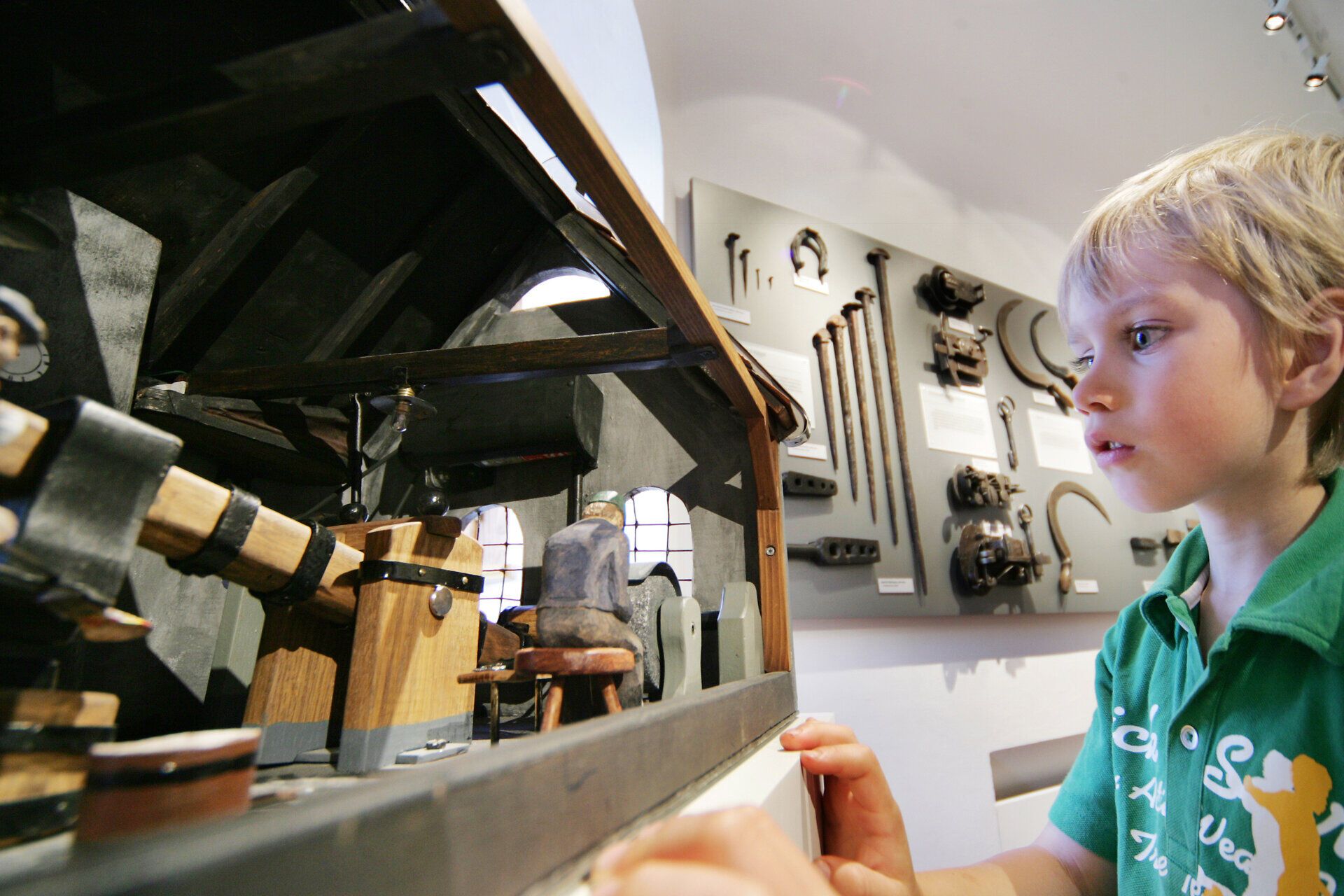 Ein kleiner Junge betrachtet fasziniert ein detailreiches Modell einer Schmiede, das die Kunst des Eisenhandwerks lebendig werden lässt. Die Atmosphäre ist geprägt von Neugier und Staunen, während die Geschichte des Schmiedezentrums in jedem Detail des Modells spürbar wird.