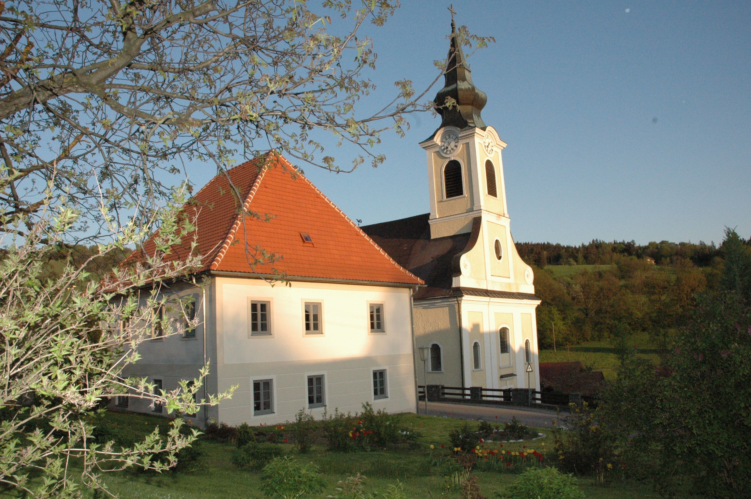 Kirche mit Pfarrhof in ländlicher Umgebung, umgeben von Bäumen und Wiesen.