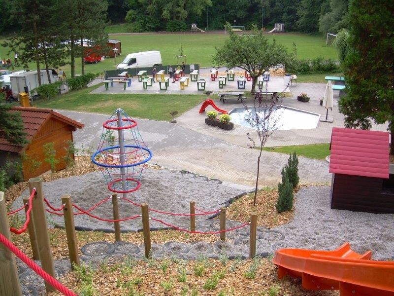 Spielplatz mit Klettergerüst, Rutsche und kleinem Pool, umgeben von Bäumen und Wiese.