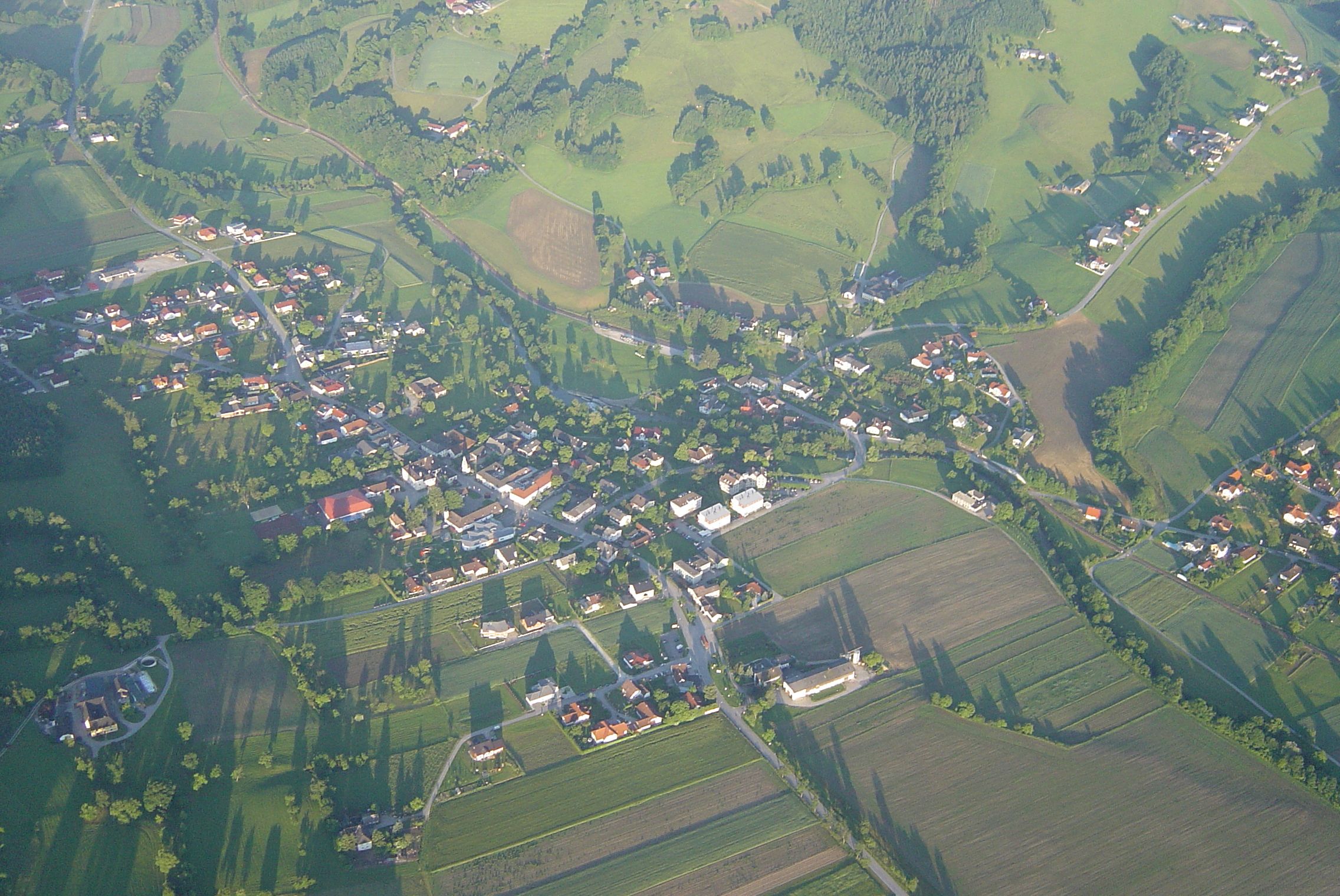 Luftaufnahme eines kleinen Dorfes mit umliegenden Feldern und Wäldern.
