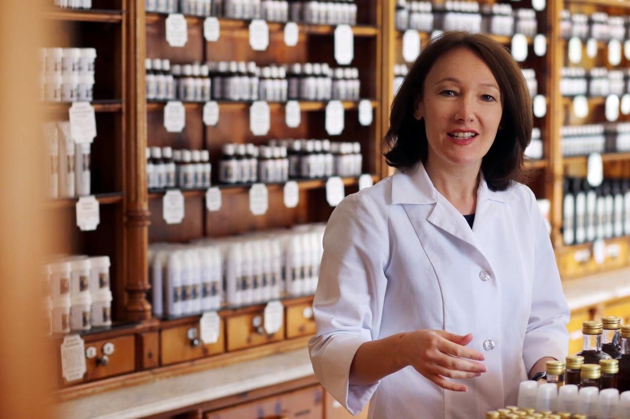 Frau in weißem Kittel in einer traditionellen Apotheke mit Regalen voller Flaschen.