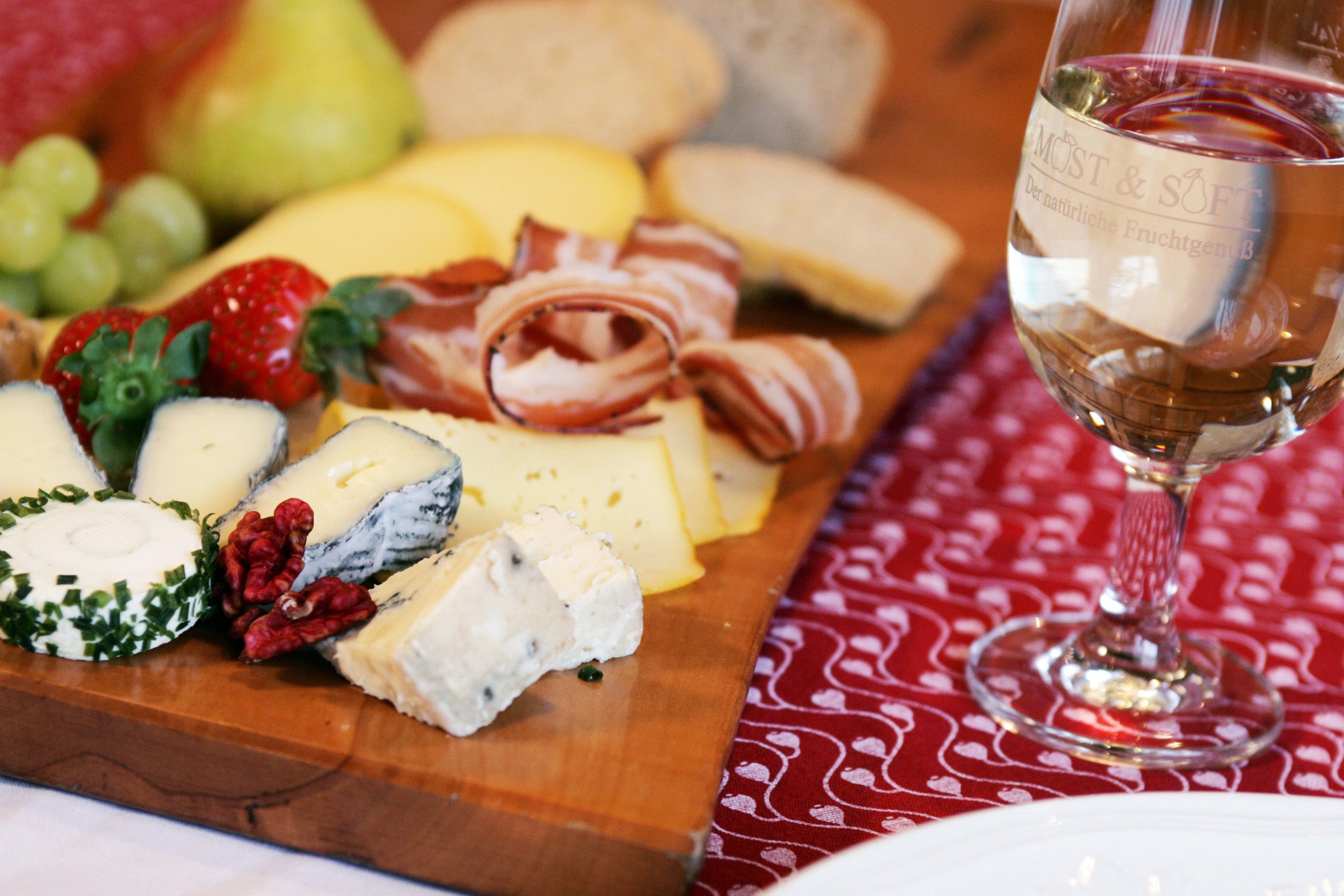 Eine Brettljause mit Käse, Schinken, Brot und Obst auf einem Holzbrett, daneben ein Glas Weißwein.
