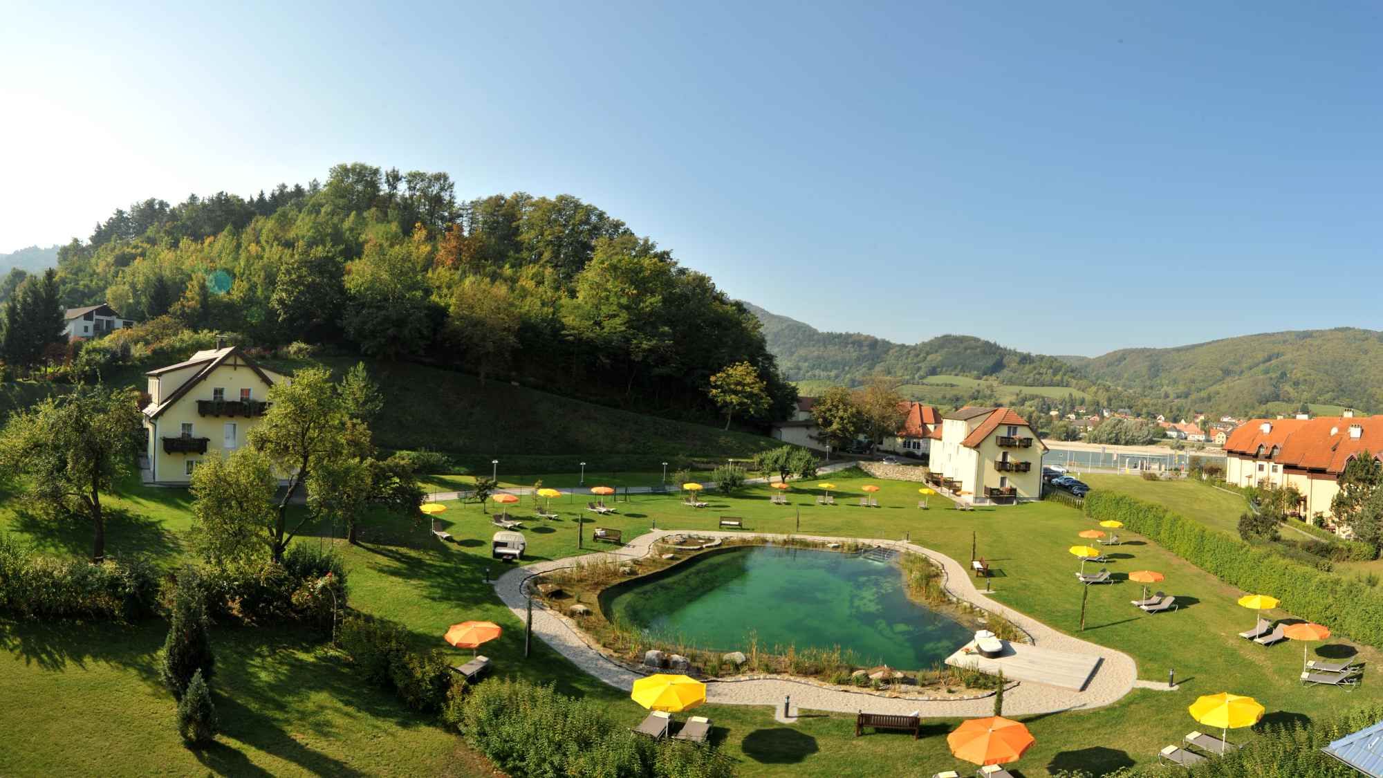 Gartenanlage mit Teich, Liegestühlen und Sonnenschirmen, umgeben von Hügeln und Häusern.