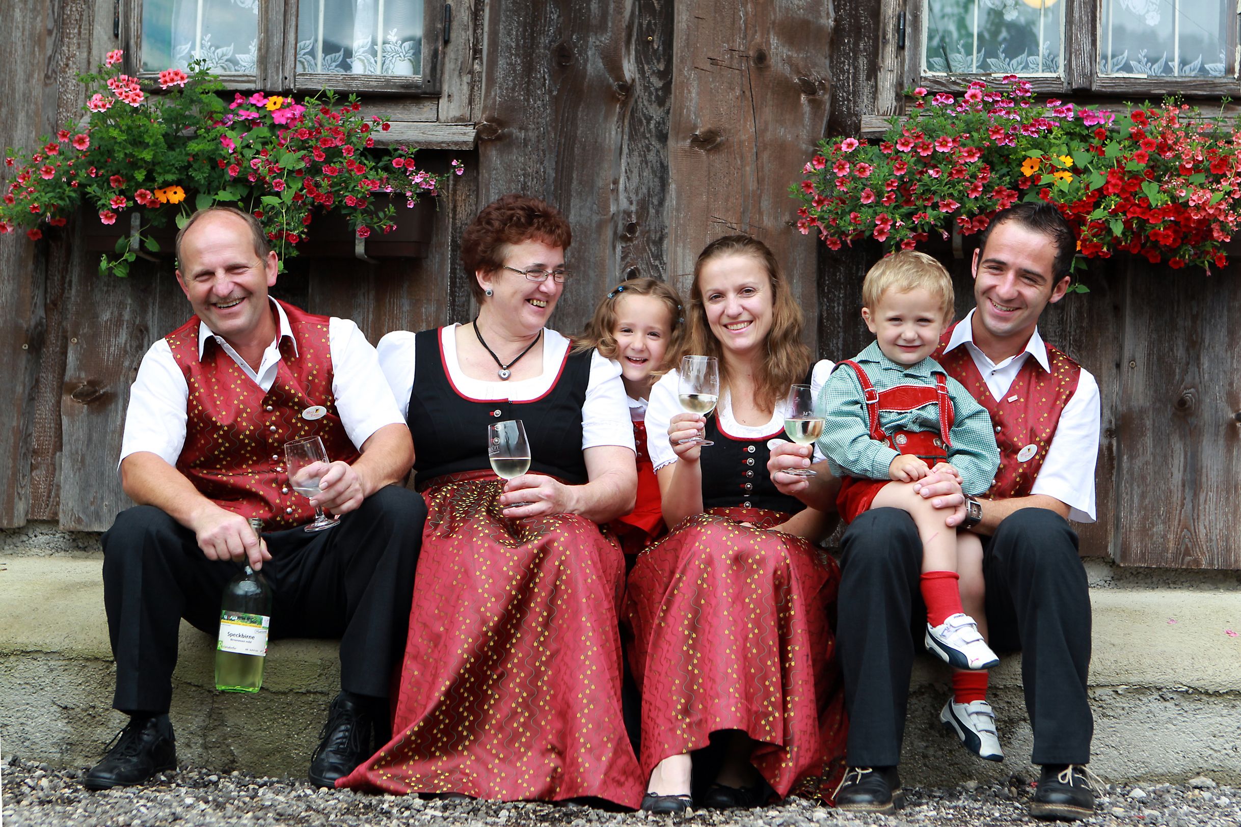 Eine Familie in traditioneller Kleidung sitzt vor einem Holzhaus mit Blumen und hält Weingläser.