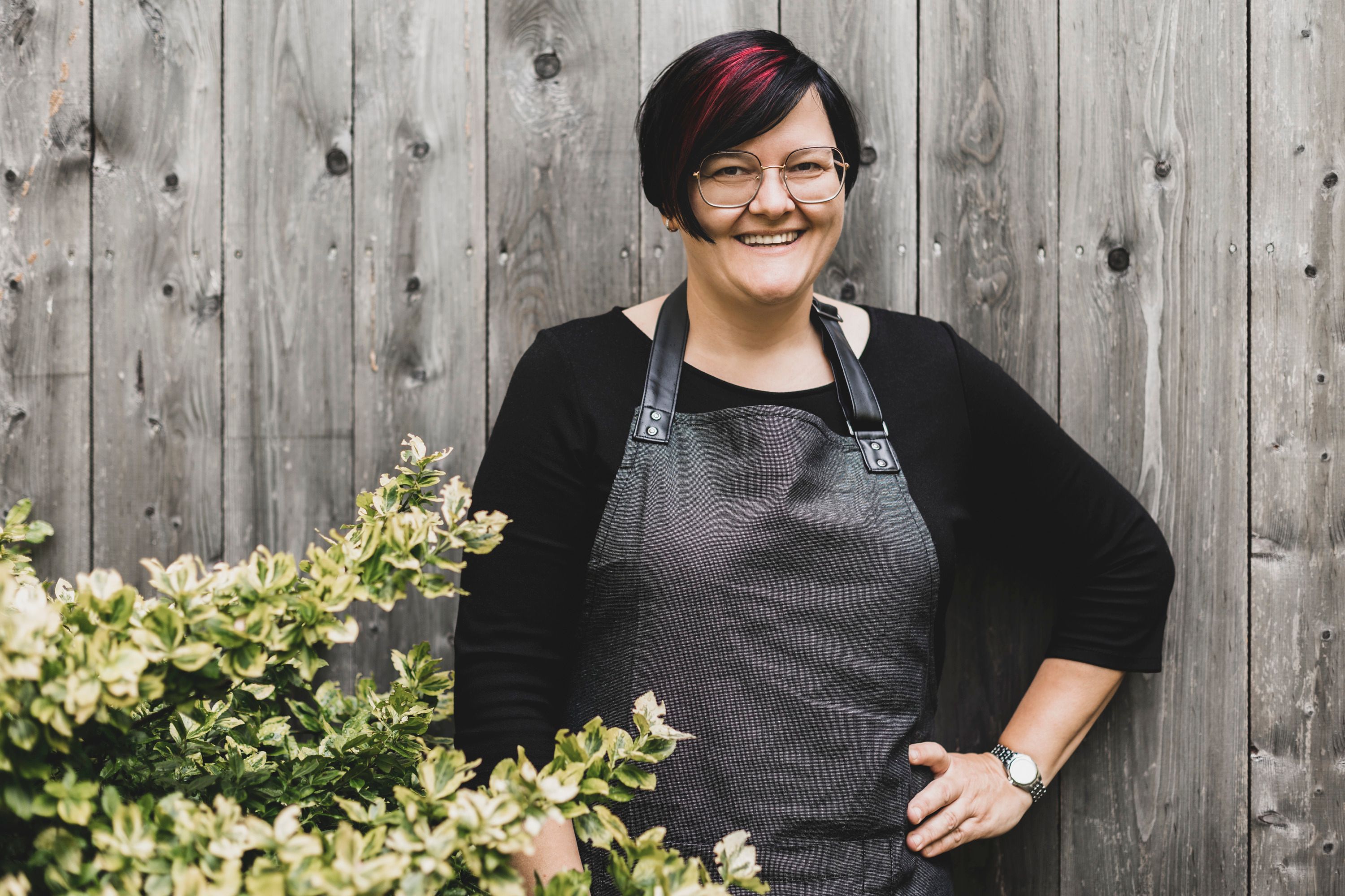 Eine Frau mit Brille und Schürze steht lächelnd vor einer Holzwand neben einem Busch.