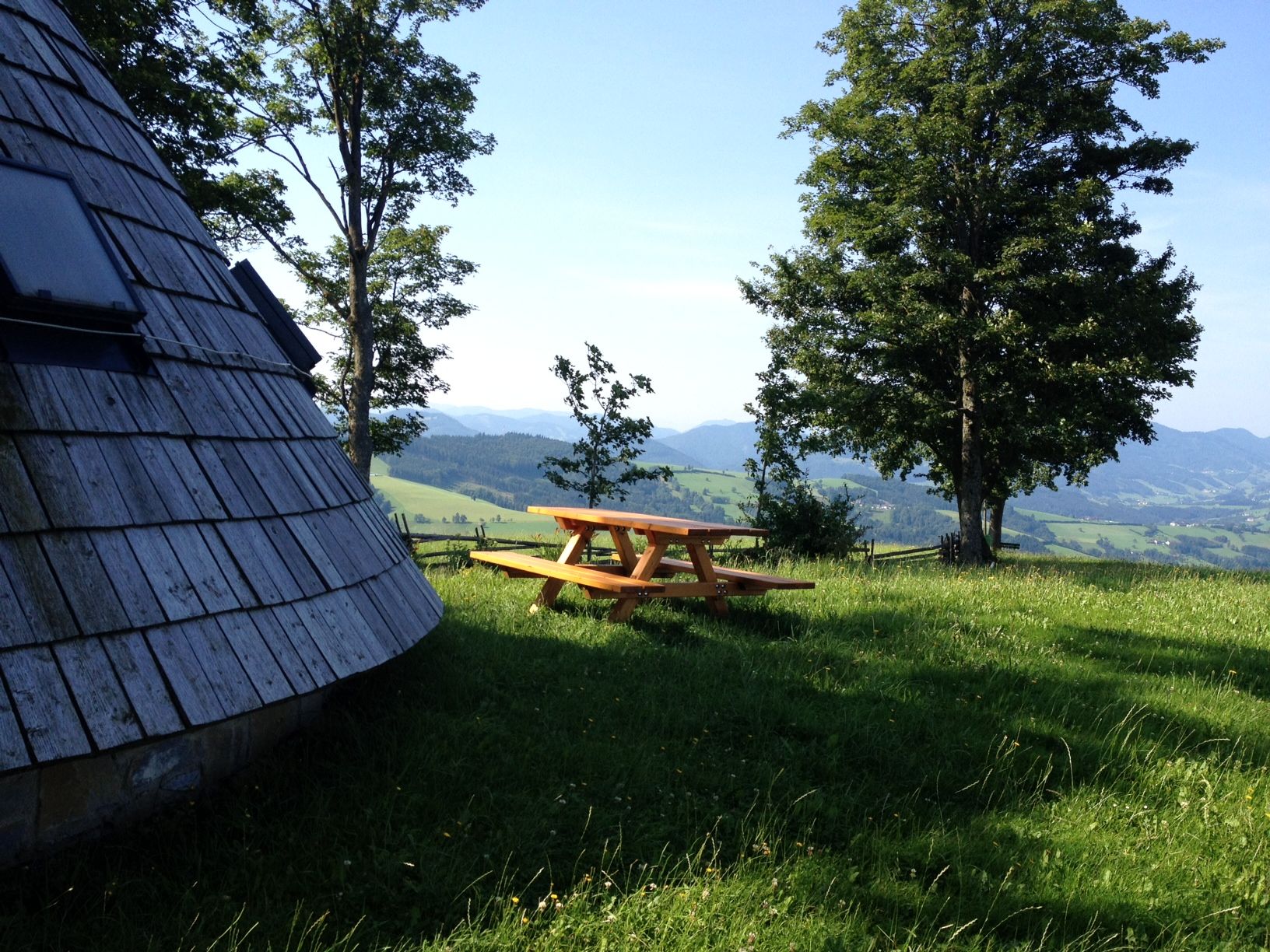 Holzhütte und Picknicktisch auf einer Wiese mit Bäumen und Bergblick.