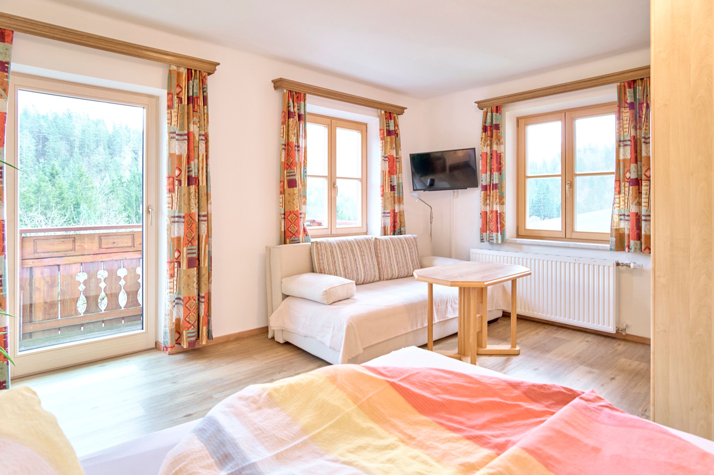 Helles Zimmer mit Sofa, Tisch, Fernseher und Balkon mit Waldblick.