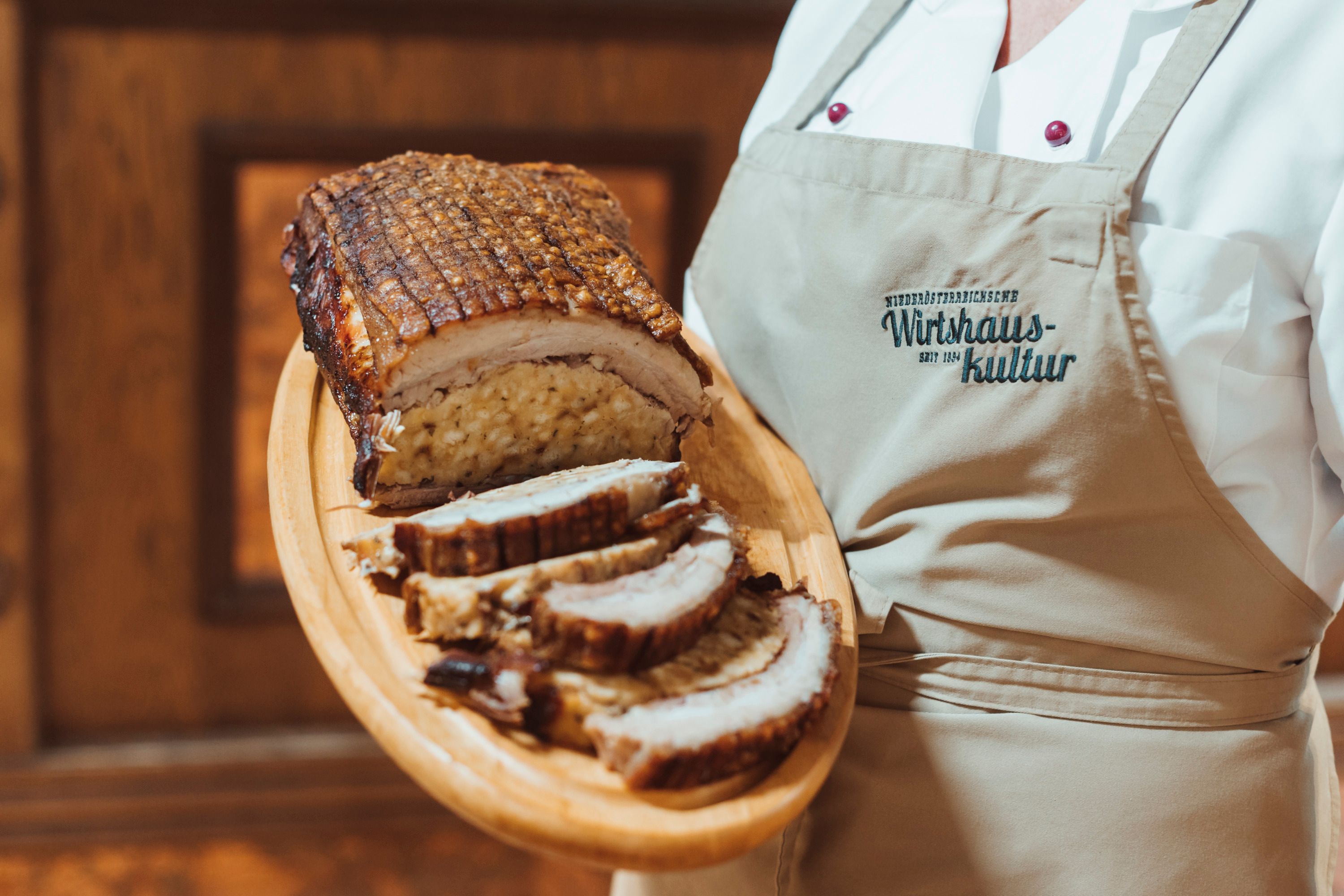Gefüllte Schweinebrust auf Holzplatte, serviert von Person in Schürze.