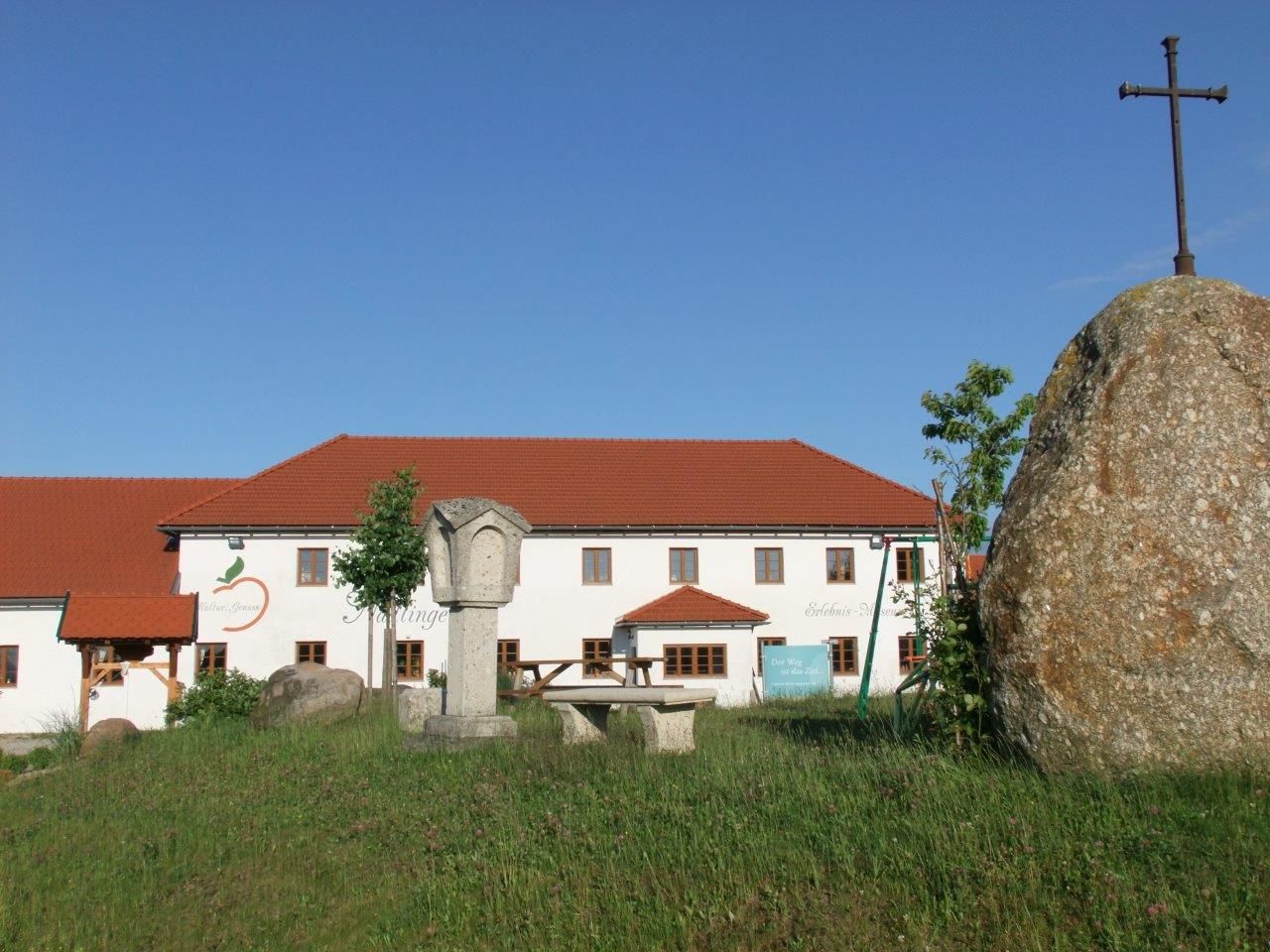 Ein Bauernhof mit rotem Dach und einem großen Stein mit Kreuz im Vordergrund.