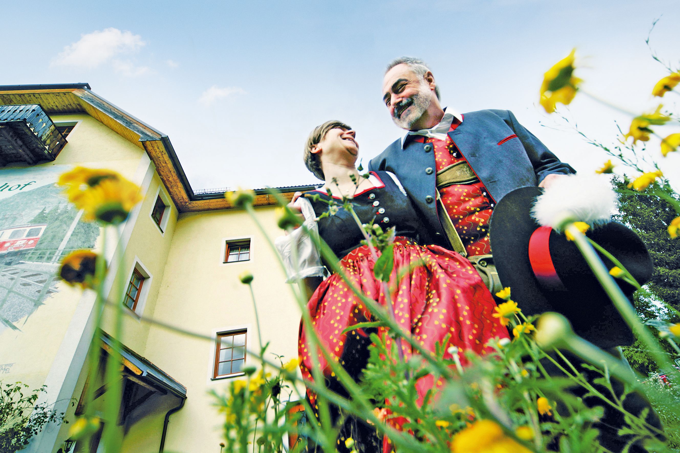 Ein Paar in traditioneller Tracht steht vor einem gelben Gebäude, umgeben von Blumen, und lächelt sich an.