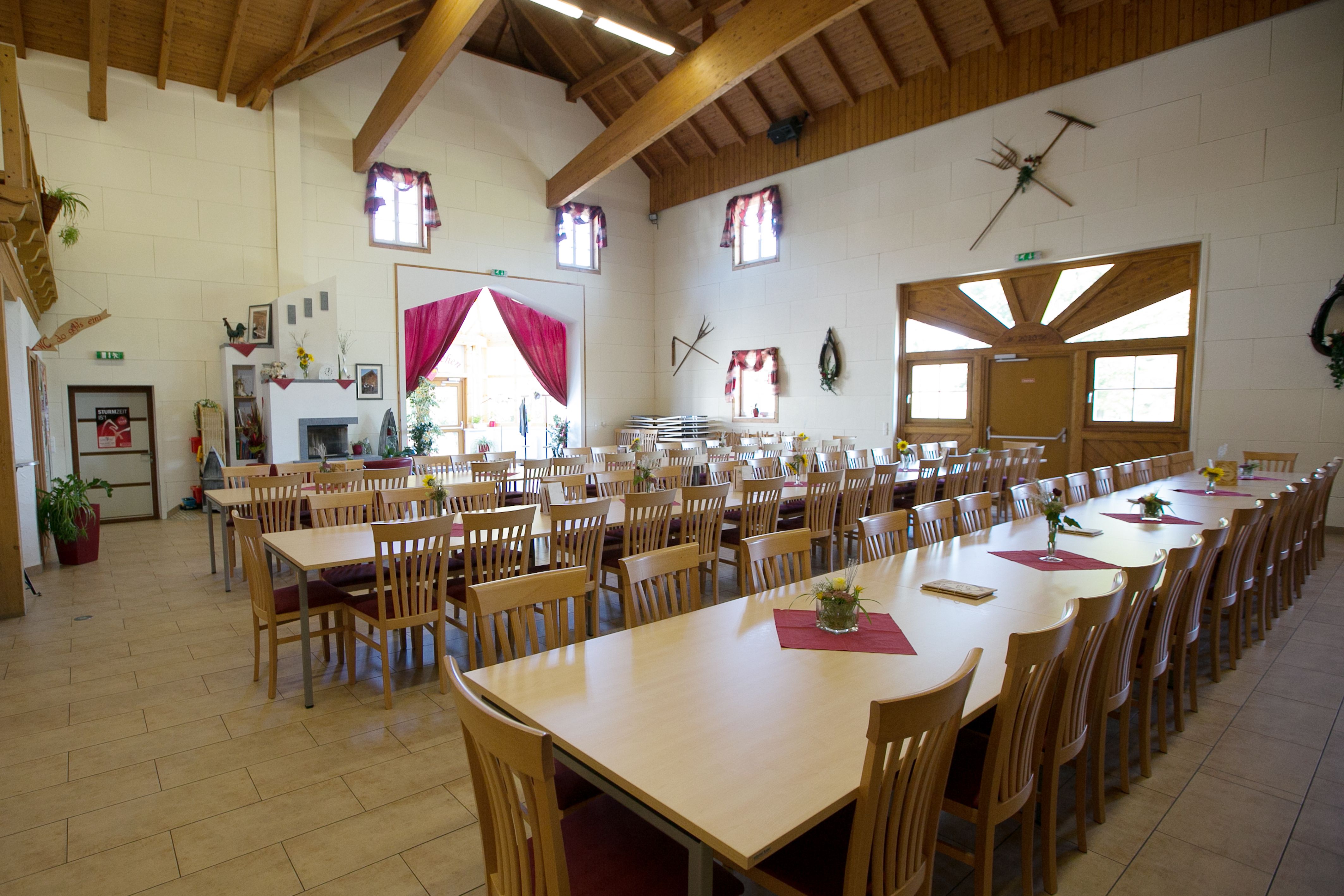 Ein großer, heller Raum mit langen Holztischen und Stühlen, dekoriert mit Blumen und roten Tischläufern, unter einem Holzdach.
