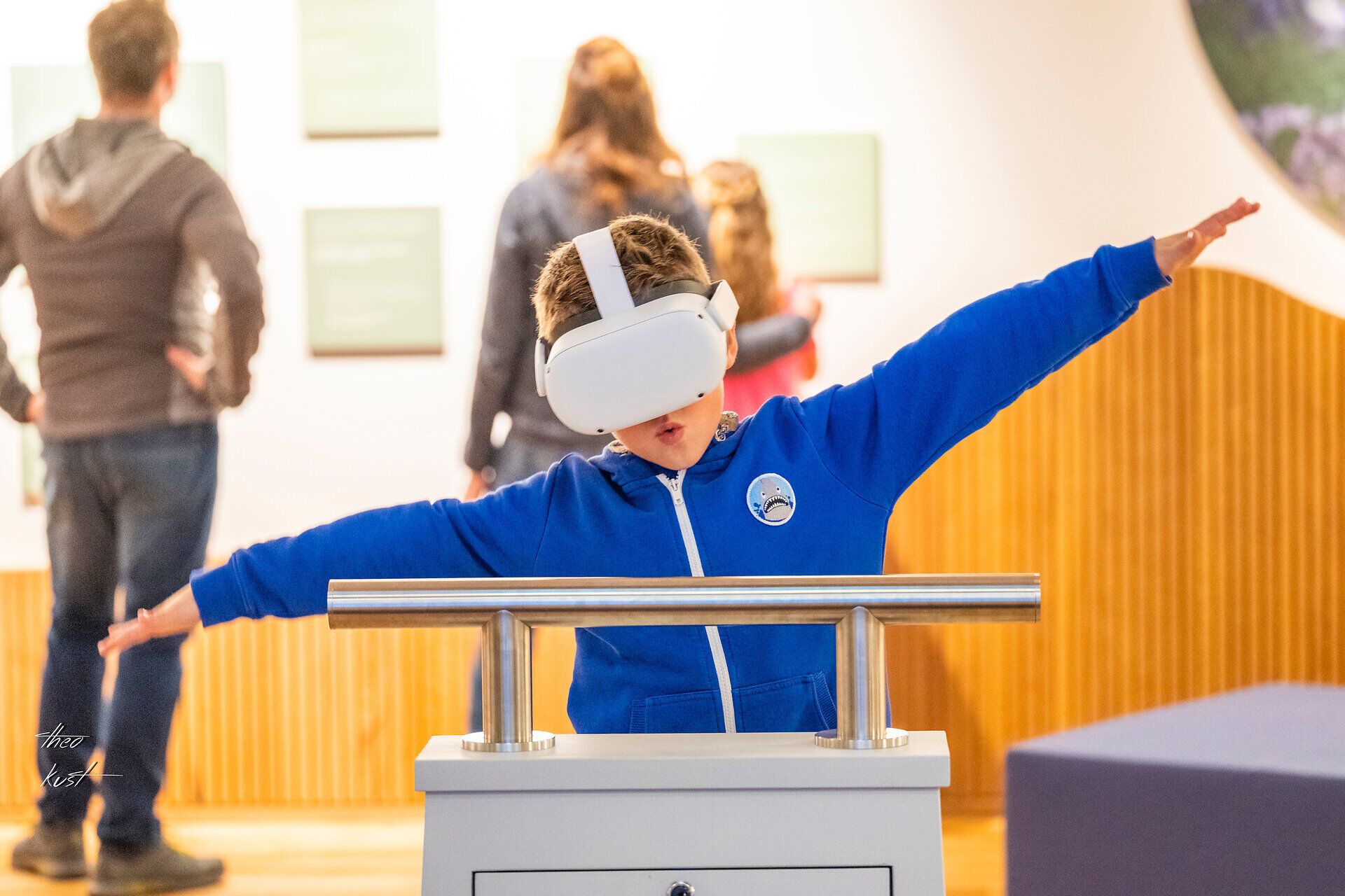 Ein Junge taucht in die faszinierende Welt der virtuellen Realität ein und erlebt Abenteuer, während seine Familie im Hintergrund die Ausstellungen erkundet. Die Atmosphäre ist lebhaft und voller Entdeckungsfreude, perfekt für einen Familienausflug an einem regnerischen Tag.