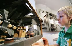 Ein kleiner Junge betrachtet fasziniert ein detailreiches Modell einer Schmiede, das die Kunst des Eisenhandwerks lebendig werden lässt. Die Atmosphäre ist geprägt von Neugier und Staunen, während die Geschichte des Schmiedezentrums in jedem Detail des Modells spürbar wird.