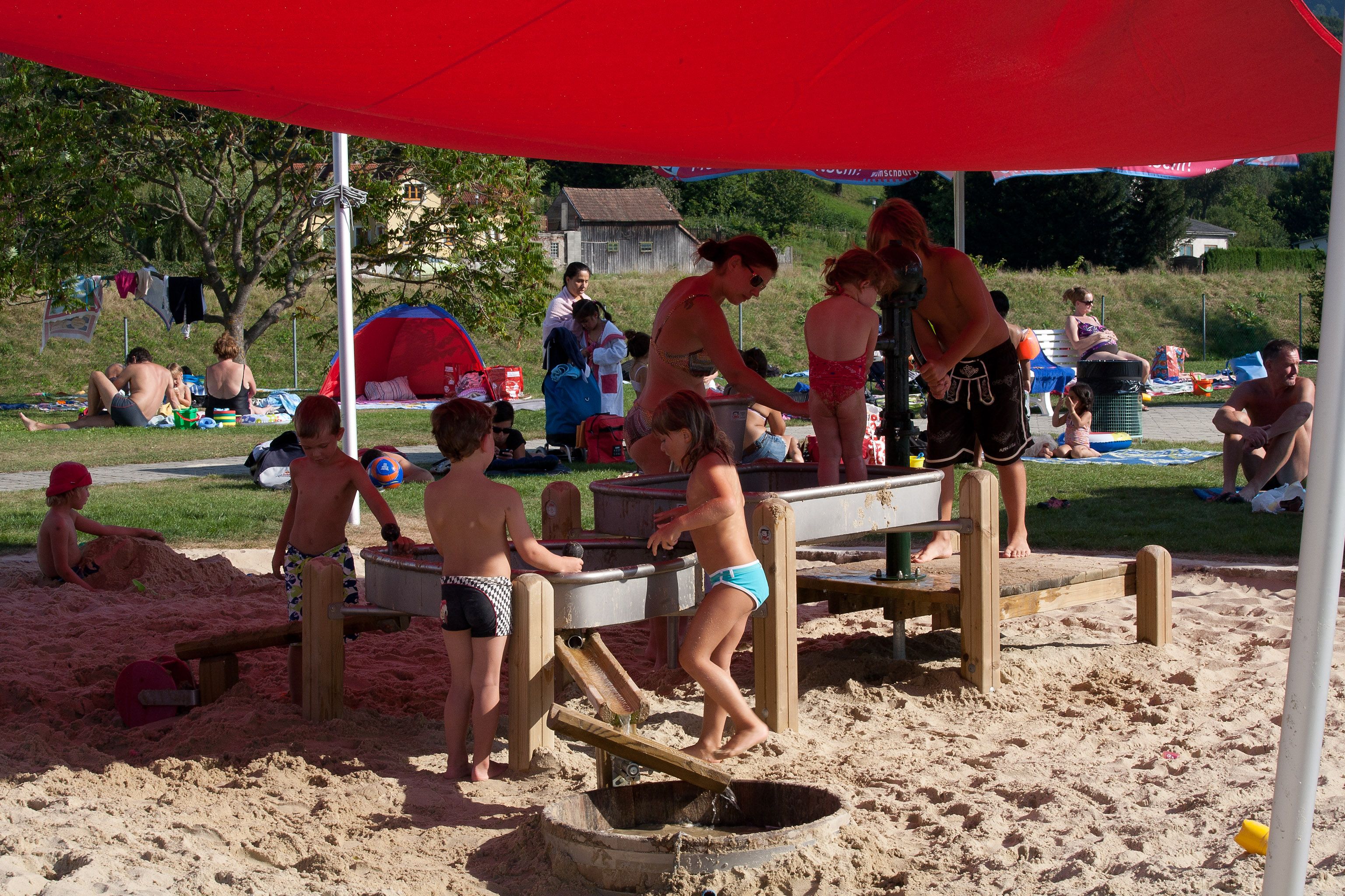 Kinder spielen im Sand unter einem roten Sonnensegel im Parkbad Wilhelmsburg.