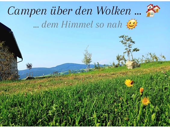 Panorama Camping am Hof Rabenberg: Wiese mit Blumen im Vordergrund, links ein Gebäude, dahinter Hügel und blauer Himmel mit dem Schriftzug „Campen über den Wolken … dem Himmel so nah“.