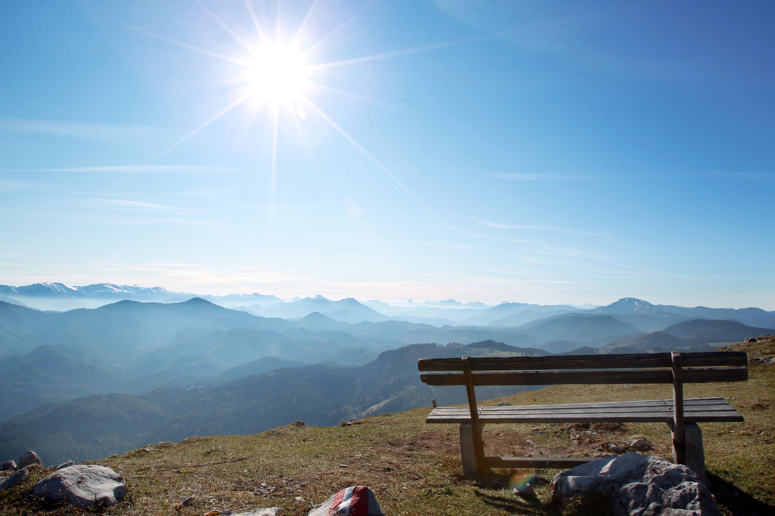 Holzbank auf einem Berg mit Blick auf eine Berglandschaft und die Sonne am Himmel.