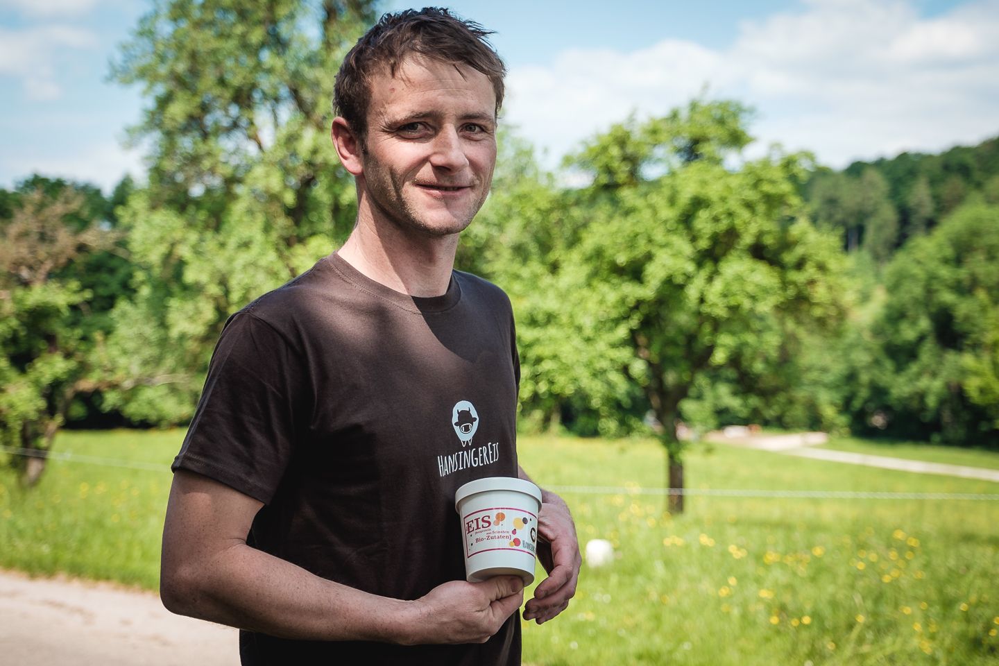 Ein Mann hält einen Becher Bioeis in einer grünen Landschaft.