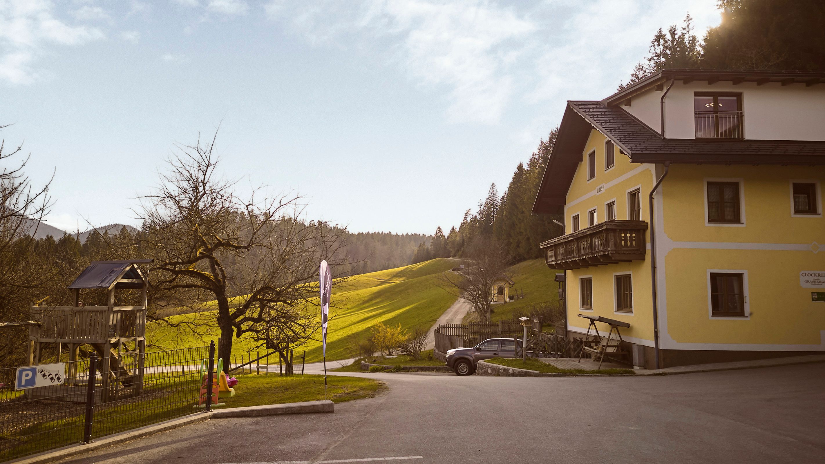 Ein gelbes Bauernhaus mit Balkon steht neben einem Spielplatz und einer grünen Wiese. Ein Auto ist auf dem Parkplatz geparkt.