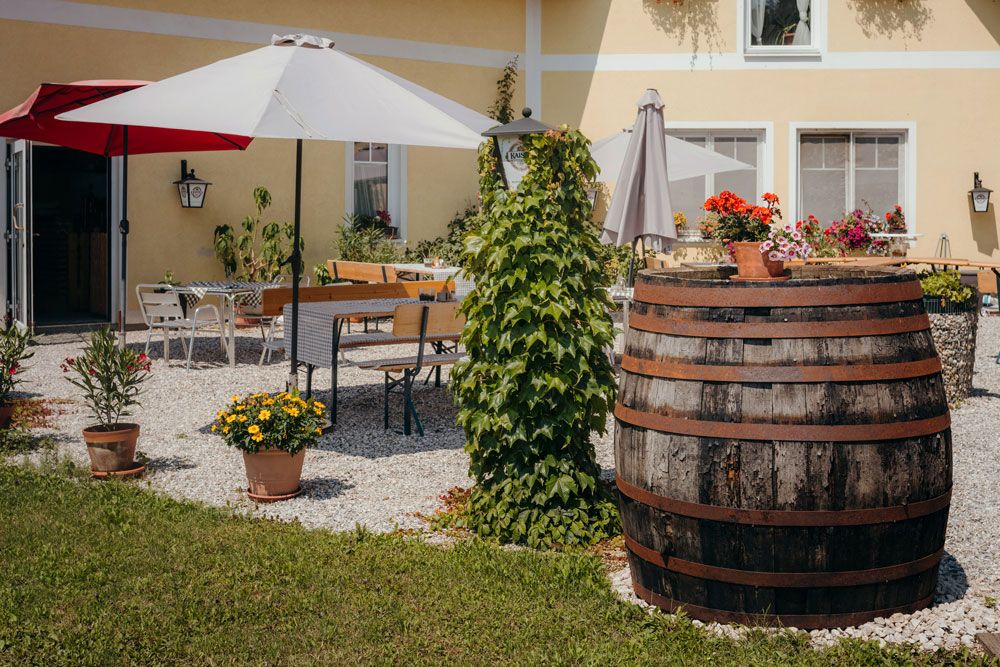 Ein gemütlicher Gastgarten mit Sonnenschirmen, Holztischen und einem großen Holzfass, umgeben von Pflanzen und Blumen.