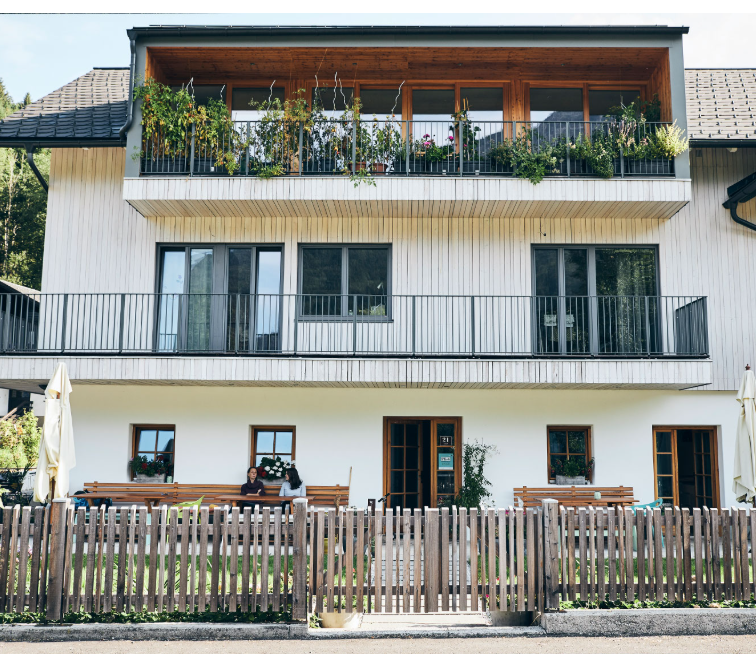Ein modernes dreistöckiges Haus mit Holzbalkonen und Pflanzen. Vor dem Haus sitzen zwei Personen auf einer Bank hinter einem Holzzaun.