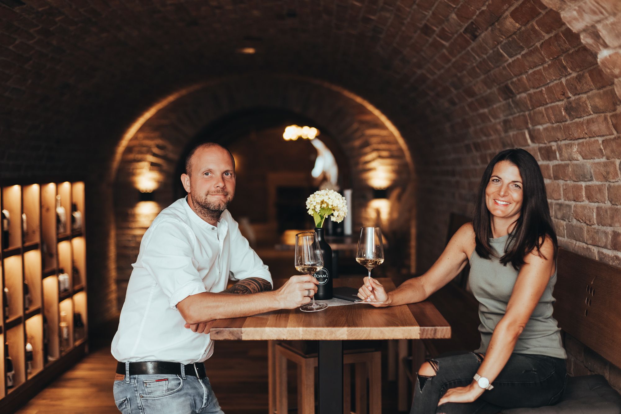 Ein Mann und eine Frau sitzen an einem Tisch in einem Weinkeller und halten Weingläser.