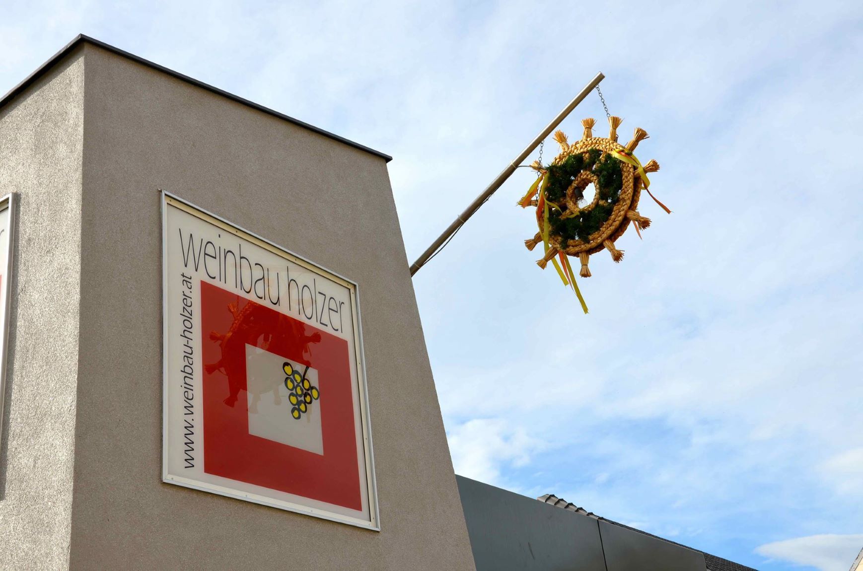 Gebäude mit Schild 'Weinbau Holzer' und einem dekorativen Kranz an einer Stange.