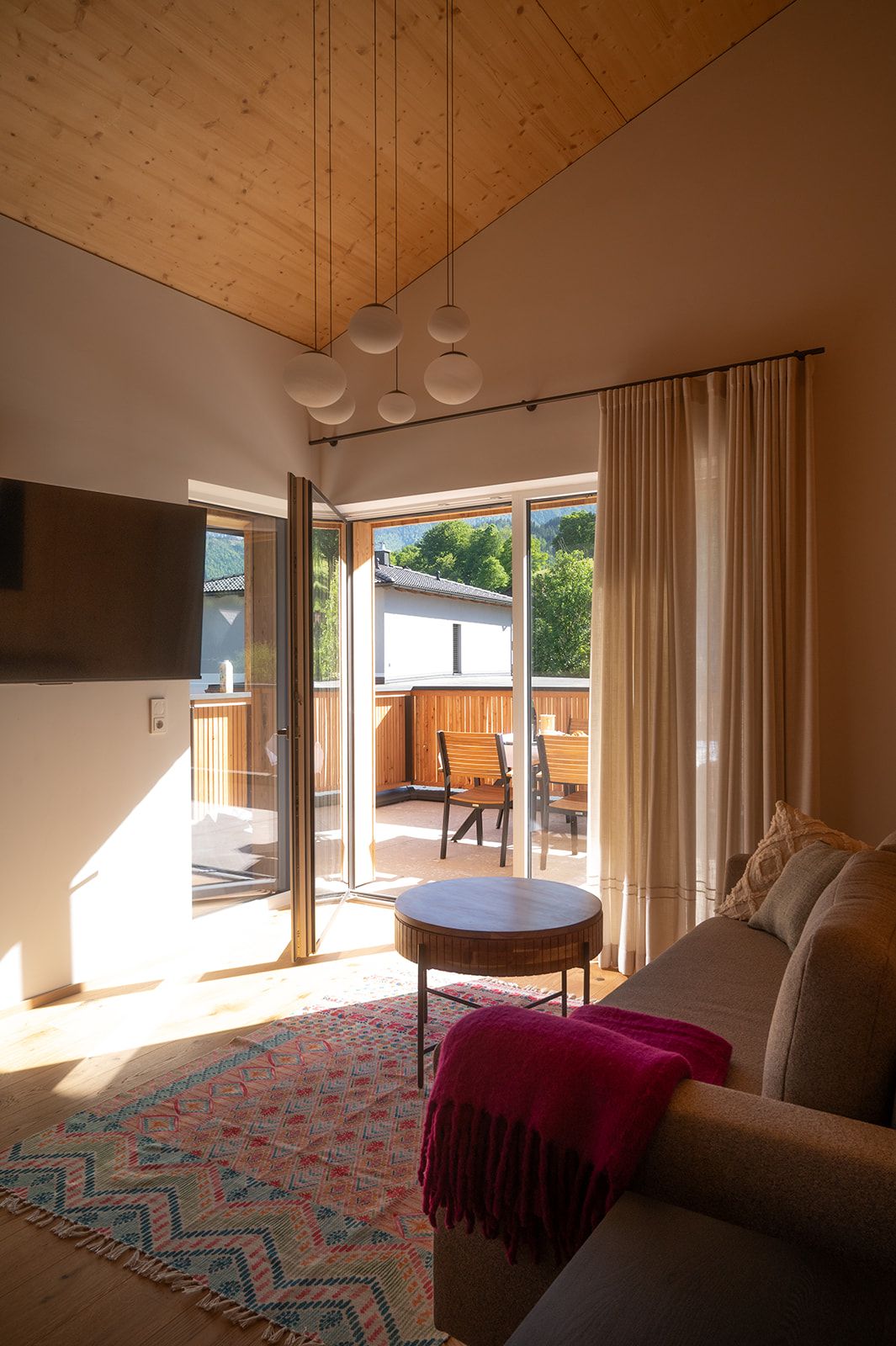 Innenansicht eines modernen Wohnzimmers mit Holzelementen, einem bunten Teppich und Zugang zu einem Balkon mit Sitzmöbeln.