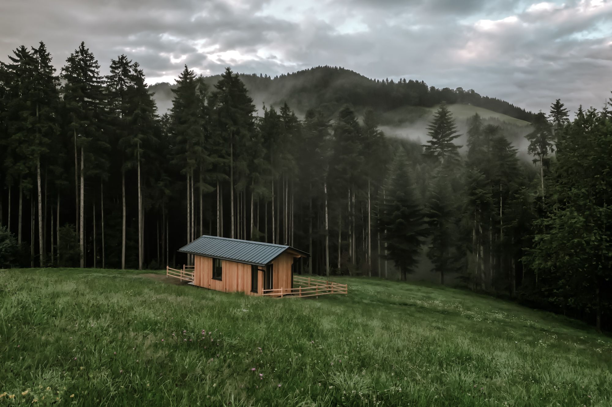 Eine kleine Holzhütte steht auf einer grünen Wiese vor einem dichten Wald mit nebligen Hügeln im Hintergrund.