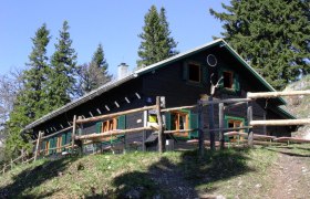 Eine Bergh&uuml;tte mit Holzzaun, umgeben von B&auml;umen, bei sonnigem Wetter.