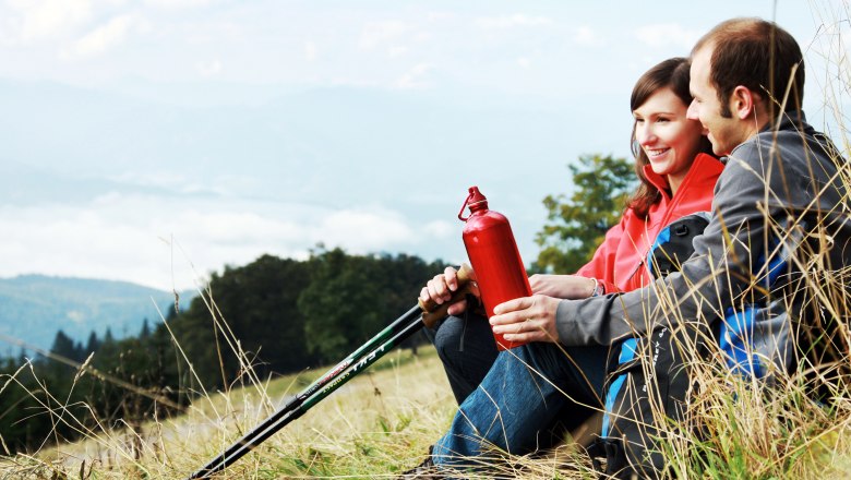 Ein Paar sitzt auf einer Wiese mit Wanderstöcken und einer roten Trinkflasche, im Hintergrund Berge und Wolken.