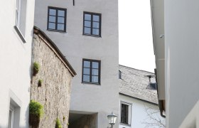 Zwei Personen gehen eine gepflasterte Gasse entlang, vorbei an einem historischen Turm mit mehreren Fenstern.