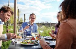 Genießen Sie die entspannte Atmosphäre eines kulinarischen Erlebnisses im Freien, umgeben von sanften Weinbergen und blühenden Pflanzen. Freunde stoßen mit Gläsern an und teilen köstliche Speisen, während die Sonne sanft auf die Tafel scheint und die Landschaft in warmes Licht taucht.