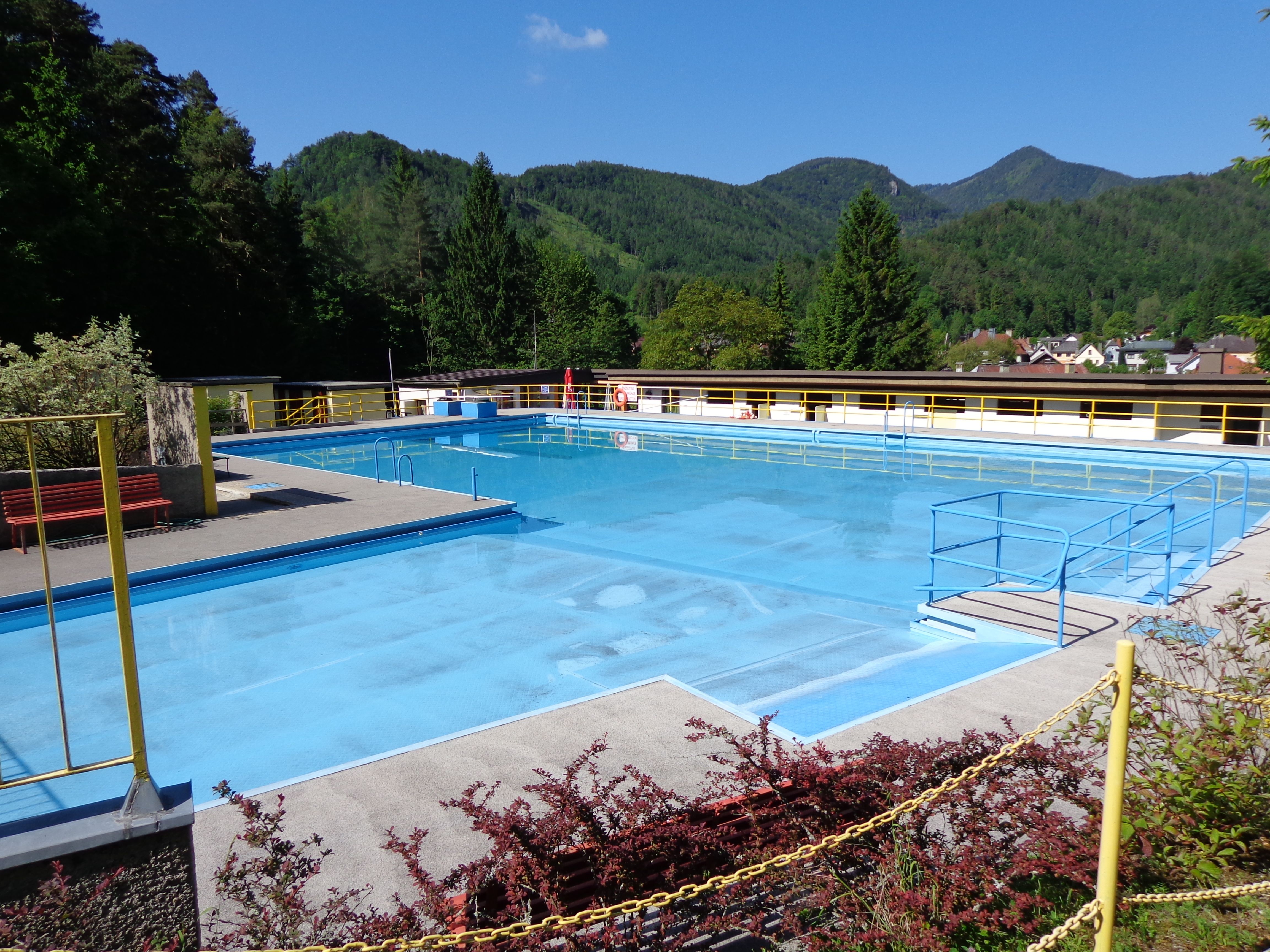 Ein leeres Freibad mit blauem Wasser, umgeben von Bergen und Bäumen.