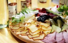 Ein Jausenbrett mit Käse, Schinken, Gurken, Nüssen und Salat auf einem Holztisch.