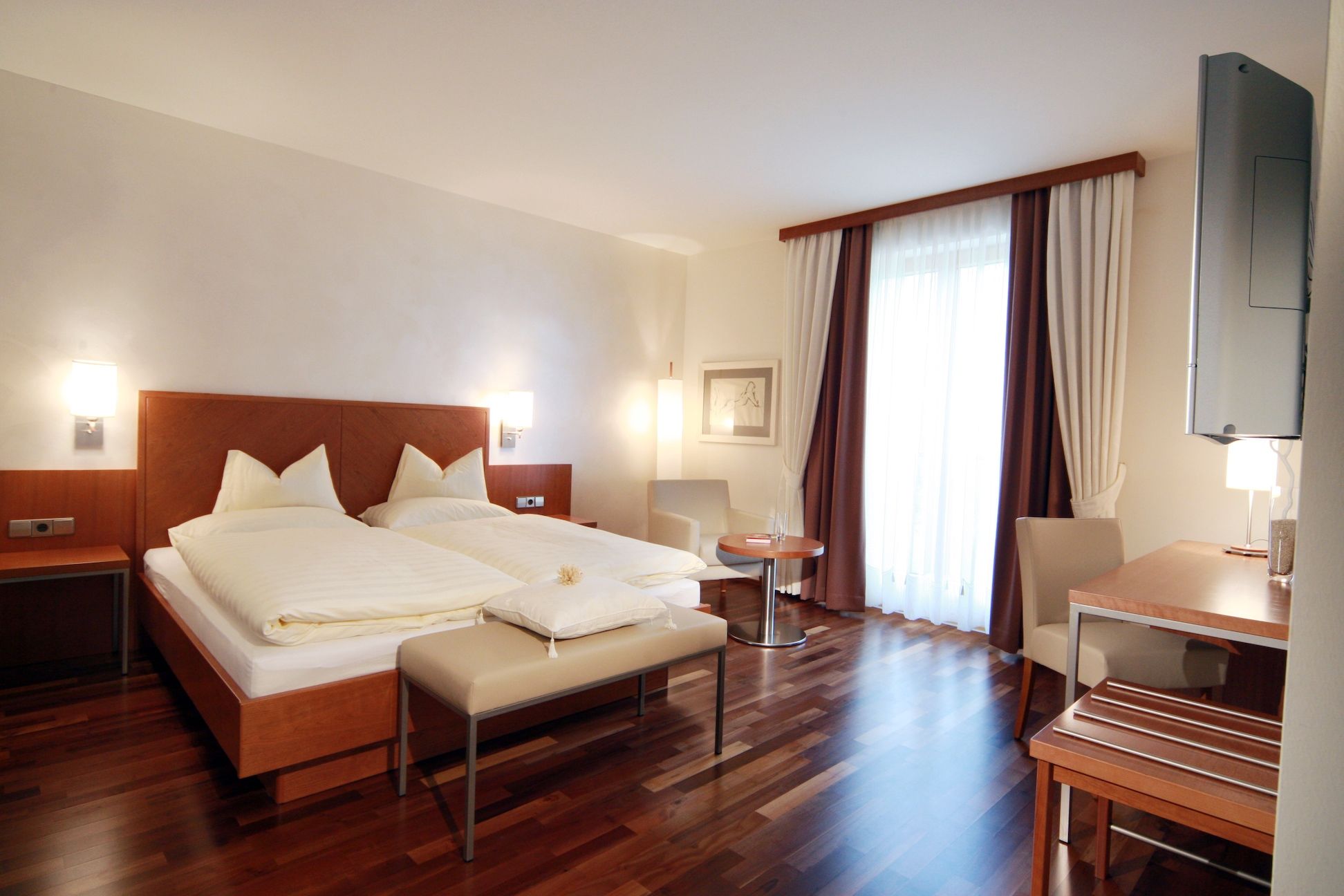 Ein modernes Hotelzimmer mit Doppelbett, Holzboden, Schreibtisch und großem Fenster mit Vorhängen.