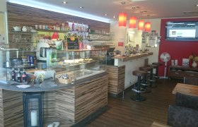 Innenansicht eines Cafés mit Theke, Barhockern und roter Wand.