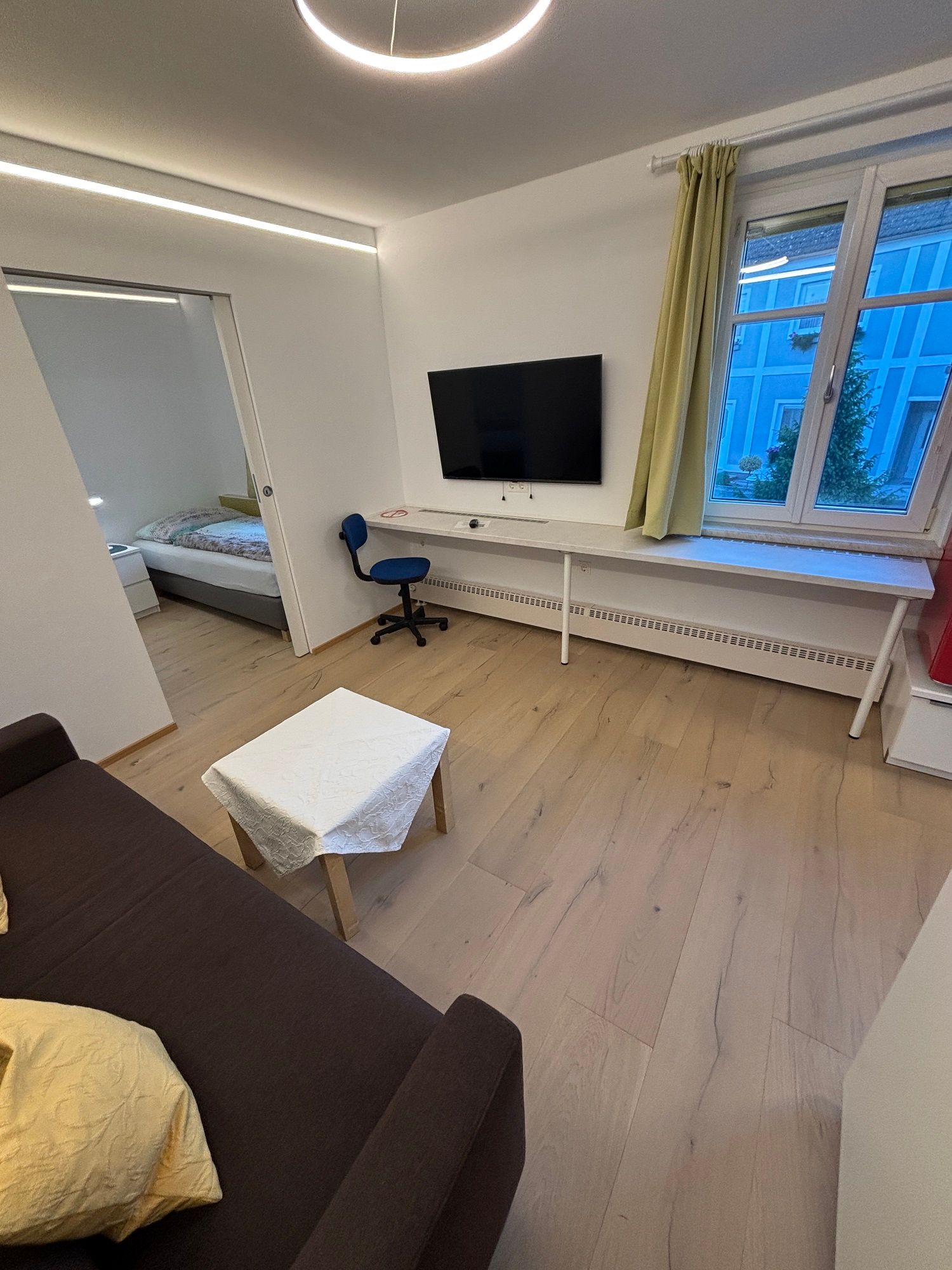 Modernes Wohnzimmer mit braunem Sofa, kleinem Tisch, Fernseher an der Wand, Schreibtisch und Stuhl. Fenster mit Blick auf ein blaues Gebäude.