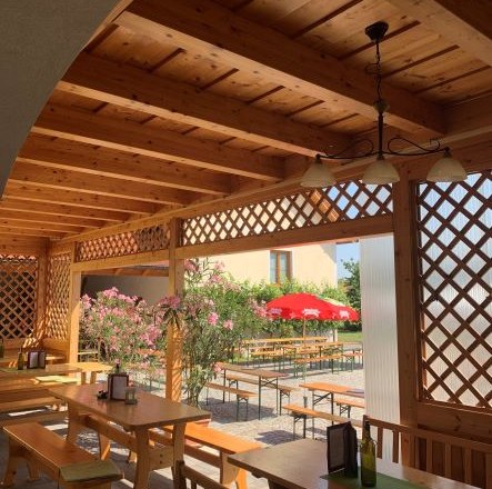 Holzpergola mit Tischen und B&auml;nken, Blick auf einen Garten mit roten Sonnenschirmen.