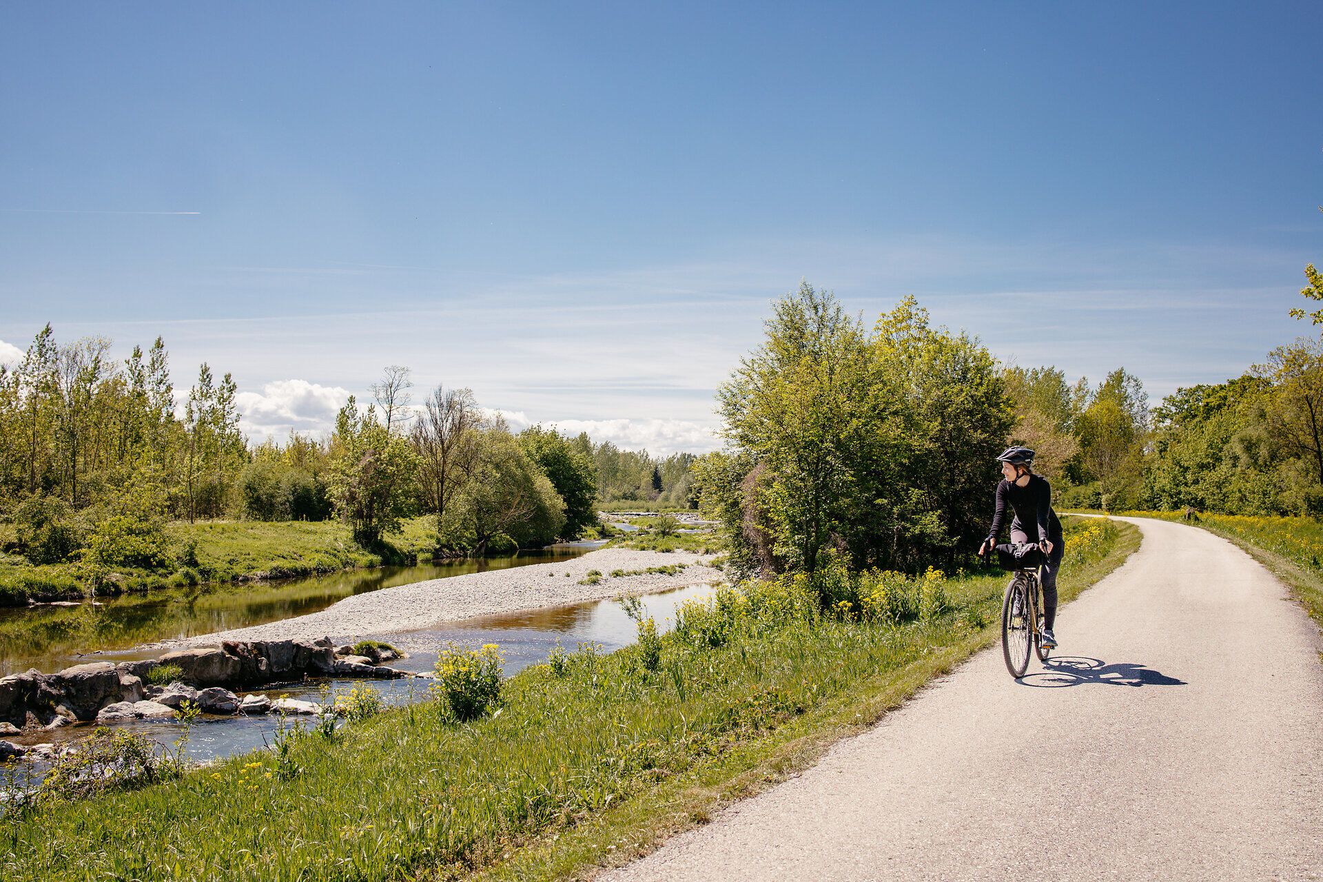 Ein malerischer Radweg schlängelt sich entlang eines sanft plätschernden Baches, umgeben von üppigem Grün und blühenden Wiesen. Die klare Luft und die strahlende Sonne laden dazu ein, die Schönheit der Natur zu genießen und die Seele baumeln zu lassen.