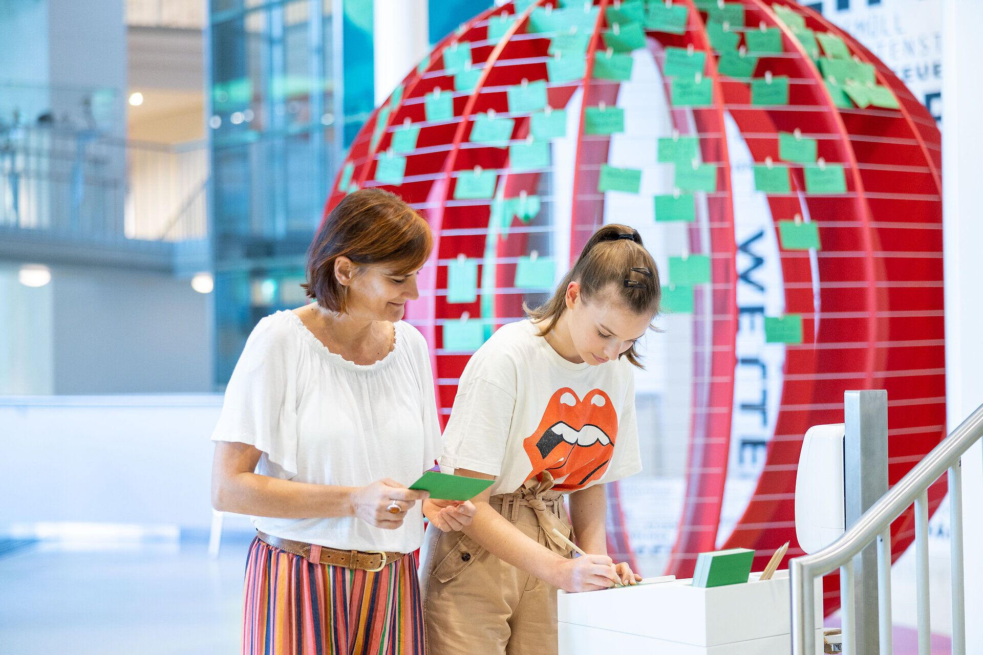 Zwei Besucherinnen stehen in einem modernen Ausstellungsraum und interagieren mit einer kreativen Installation. Die Atmosphäre ist lebhaft und inspirierend, während sie ihre Gedanken und Ideen auf bunten Karten festhalten, um gemeinsam einen Beitrag zur Umwelt zu leisten.