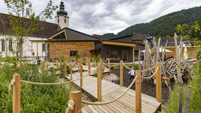 A modern building with wooden cladding and a playground, surrounded by green countryside and mountains in the background.